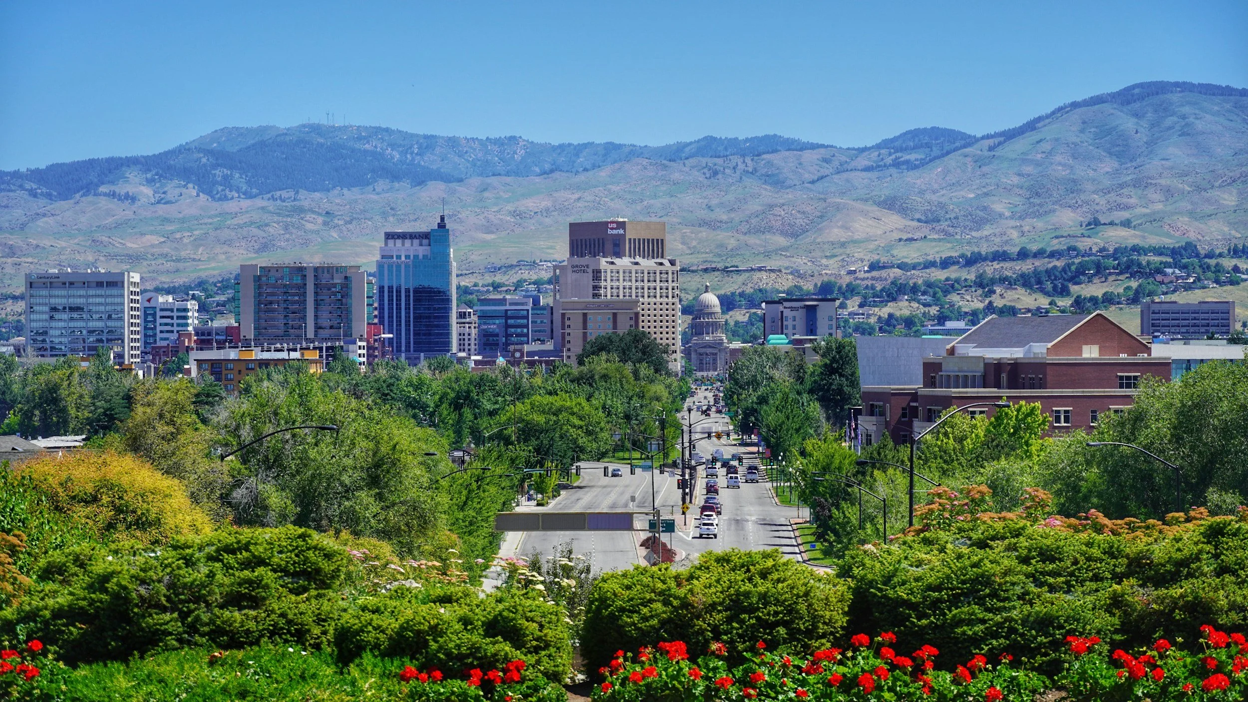 A city skyline with tall modern buildings, including one with signage for Zion's Bank and U.S. Bank, set against green hills and mountains in the background. The foreground has a street lined with trees and colorful flowers.