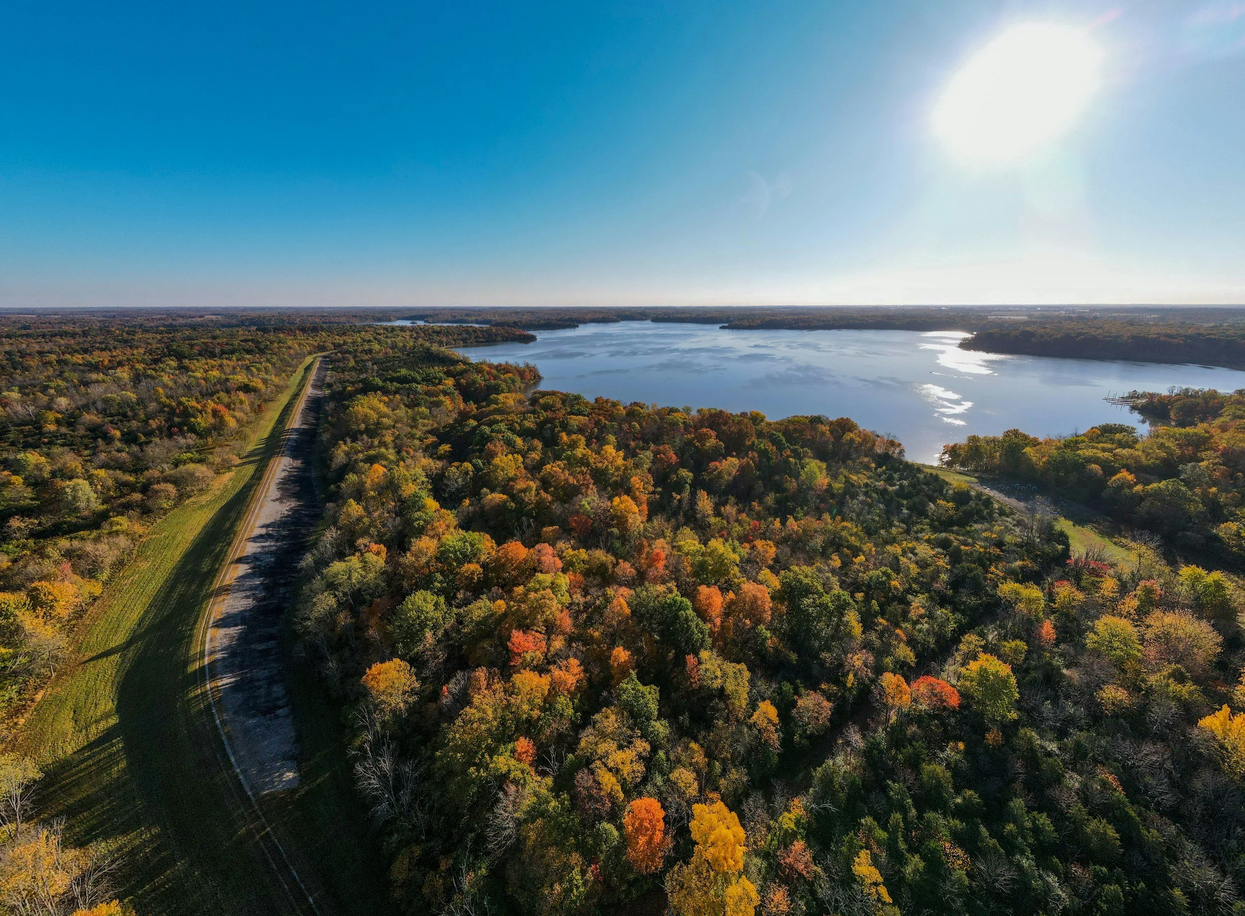 An aerial view of a large lake surrounded by dense trees with fall foliage colors, a dirt road running alongside the trees, and sunny blue sky.