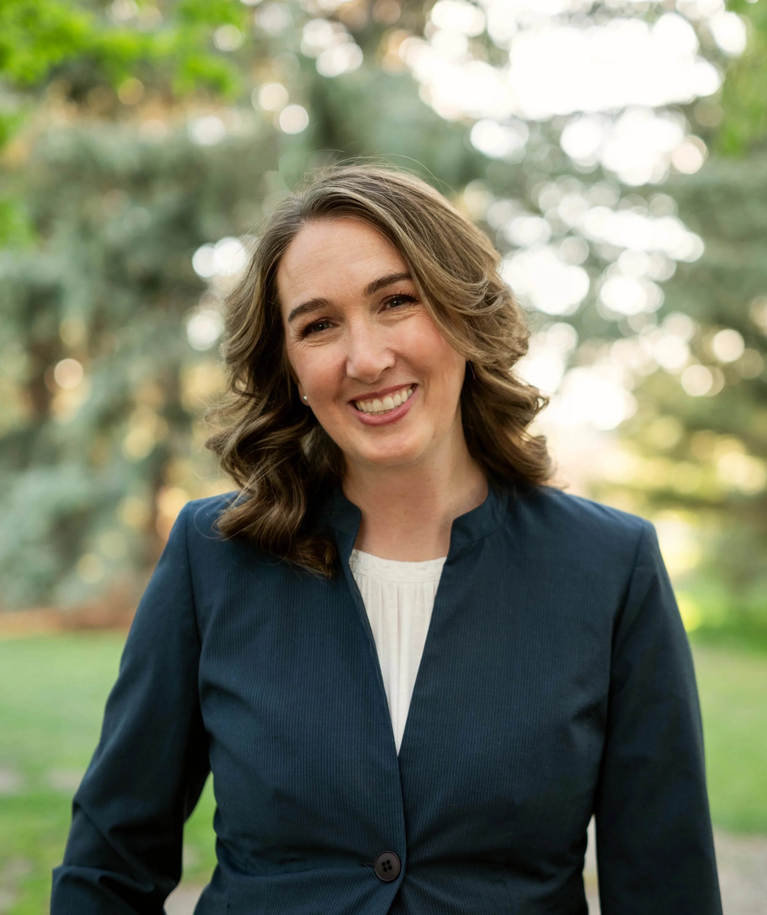 A woman with shoulder-length wavy brown hair smiling outdoors in a park with trees and sunlight in the background, wearing a dark blue blazer over a white top.