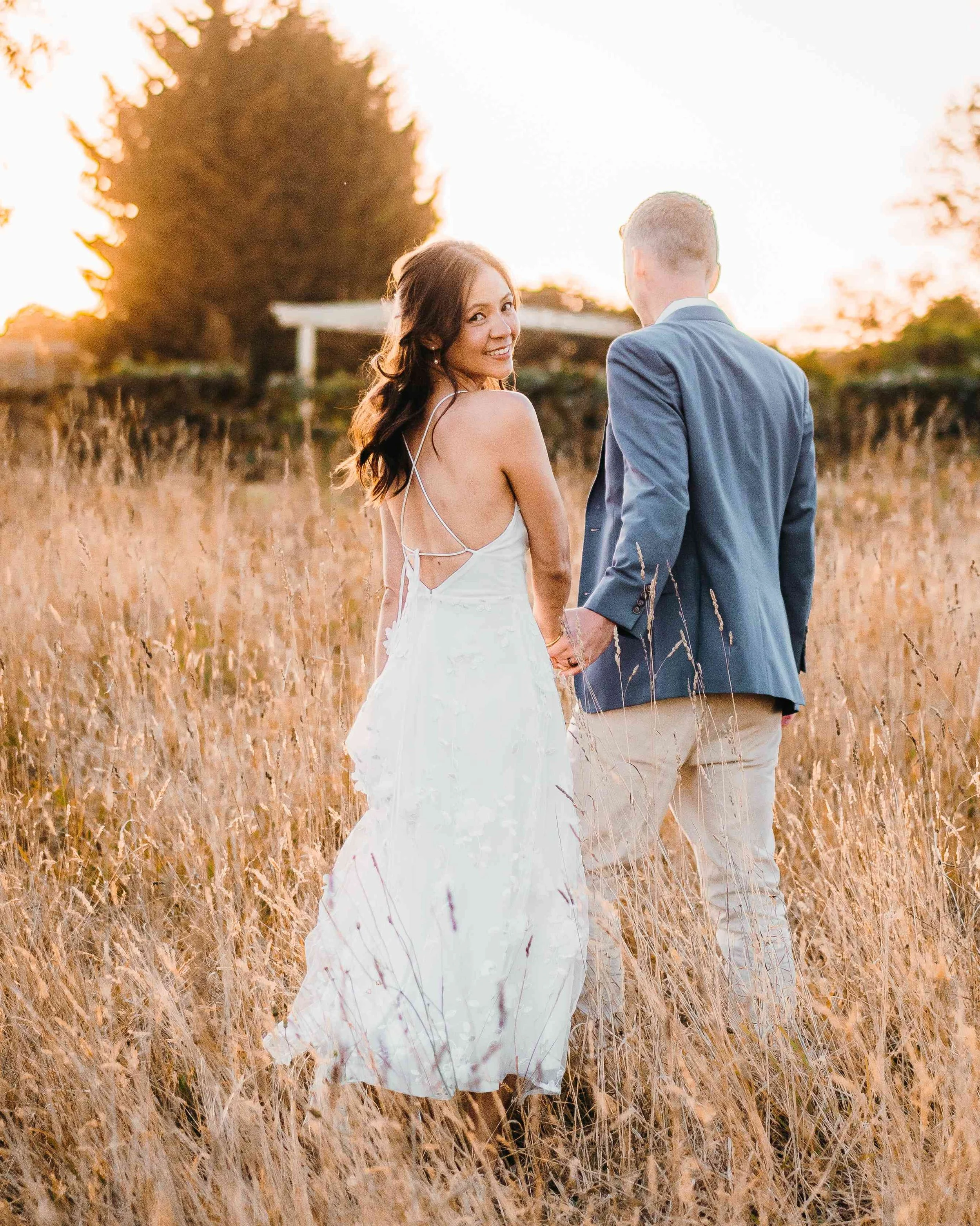 A woman in a white dress and a man in a blue jacket holding hands in a field with tall grass at sunset, smiling and looking back at the camera.