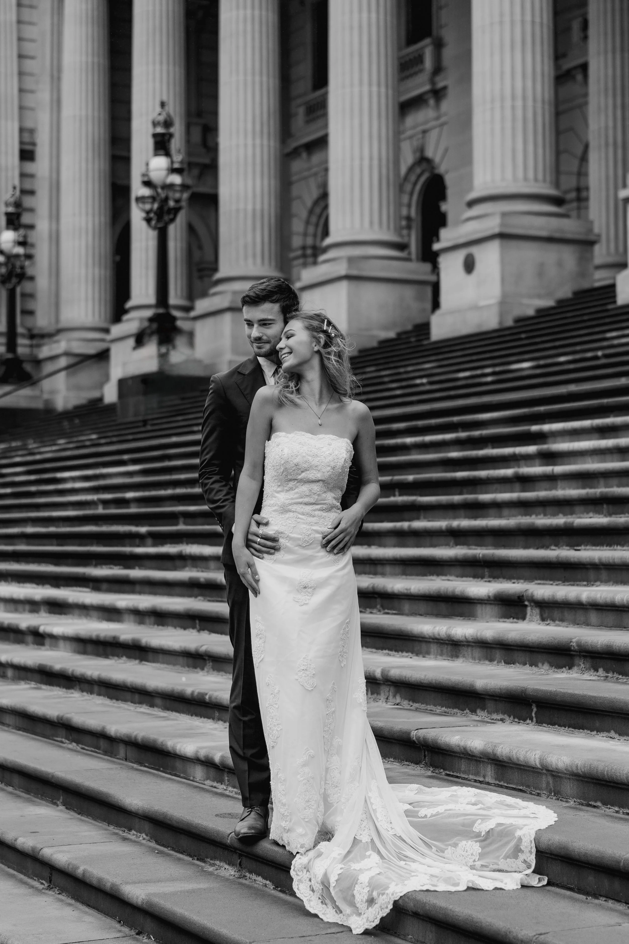 A black and white photo of a bride and groom standing on the steps of a grand building with tall columns, smiling and embracing.