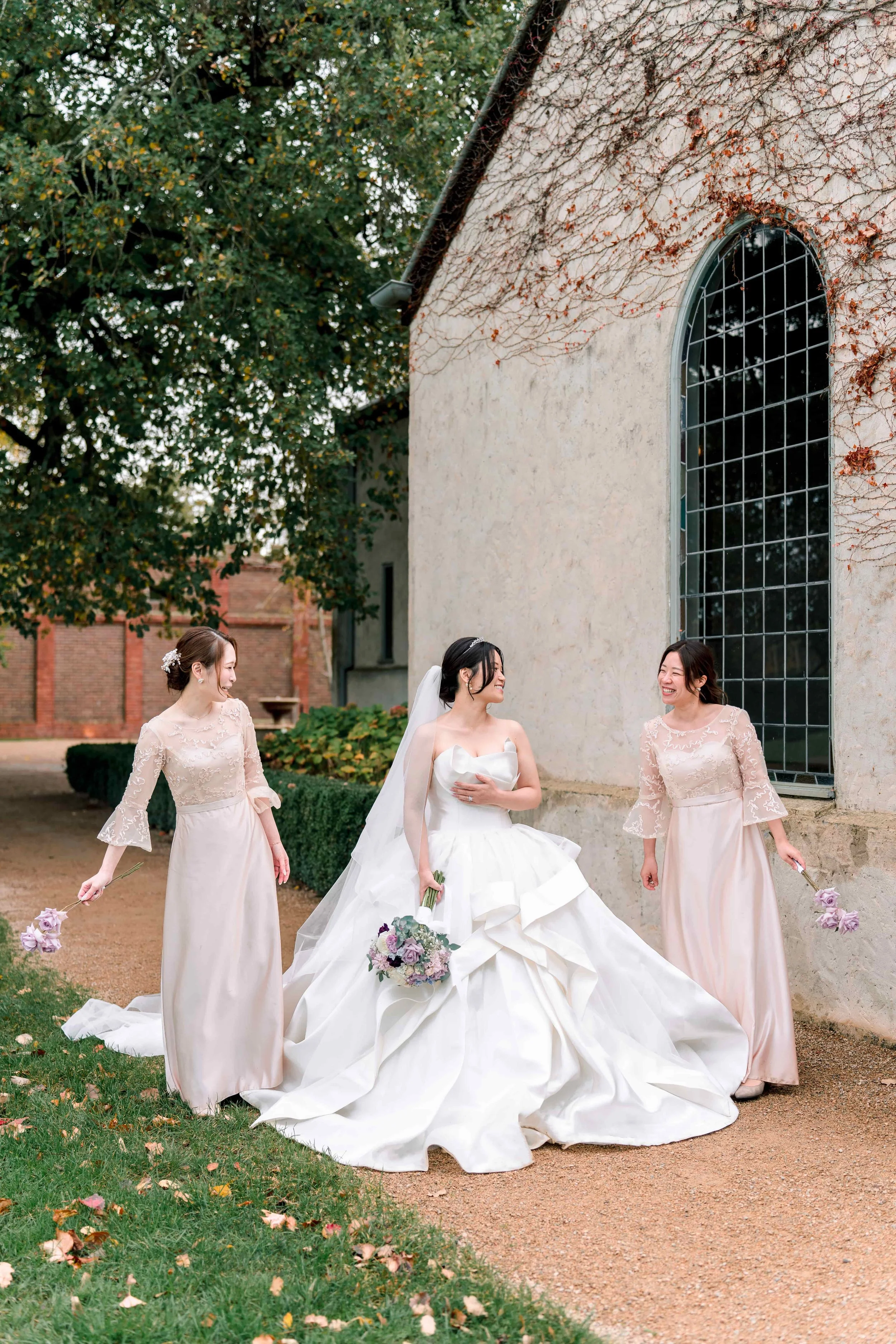 Bride in a white wedding gown holding a bouquet, surrounded by three bridesmaids in matching blush pink dresses holding small flower bouquets, standing outdoors near a building with vine-covered walls and large arched window, with trees and greenery 