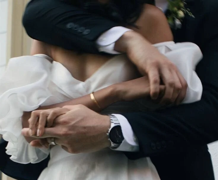 Close-up of a couple sharing a kiss, with the man holding the woman's hand, wearing a suit and watch, and the woman in a white dress.