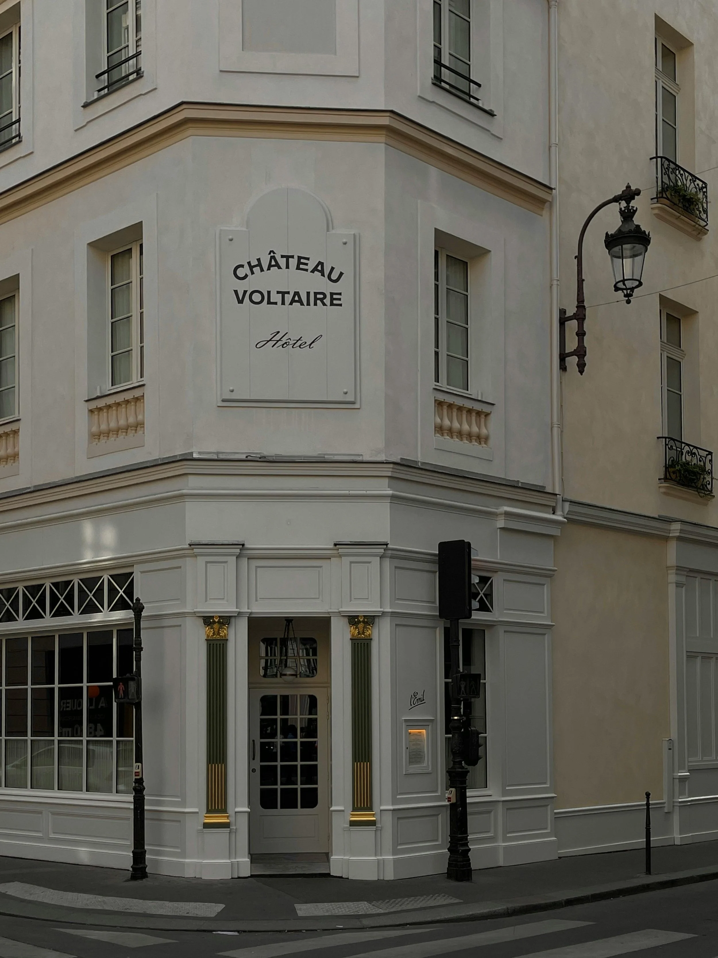A corner of a building with a sign that says 'Château Voltaire Hotel'. The building has white walls, tall windows, black wrought iron balconies, and a vintage street lamp. The street has a pedestrian crossing and traffic lights.