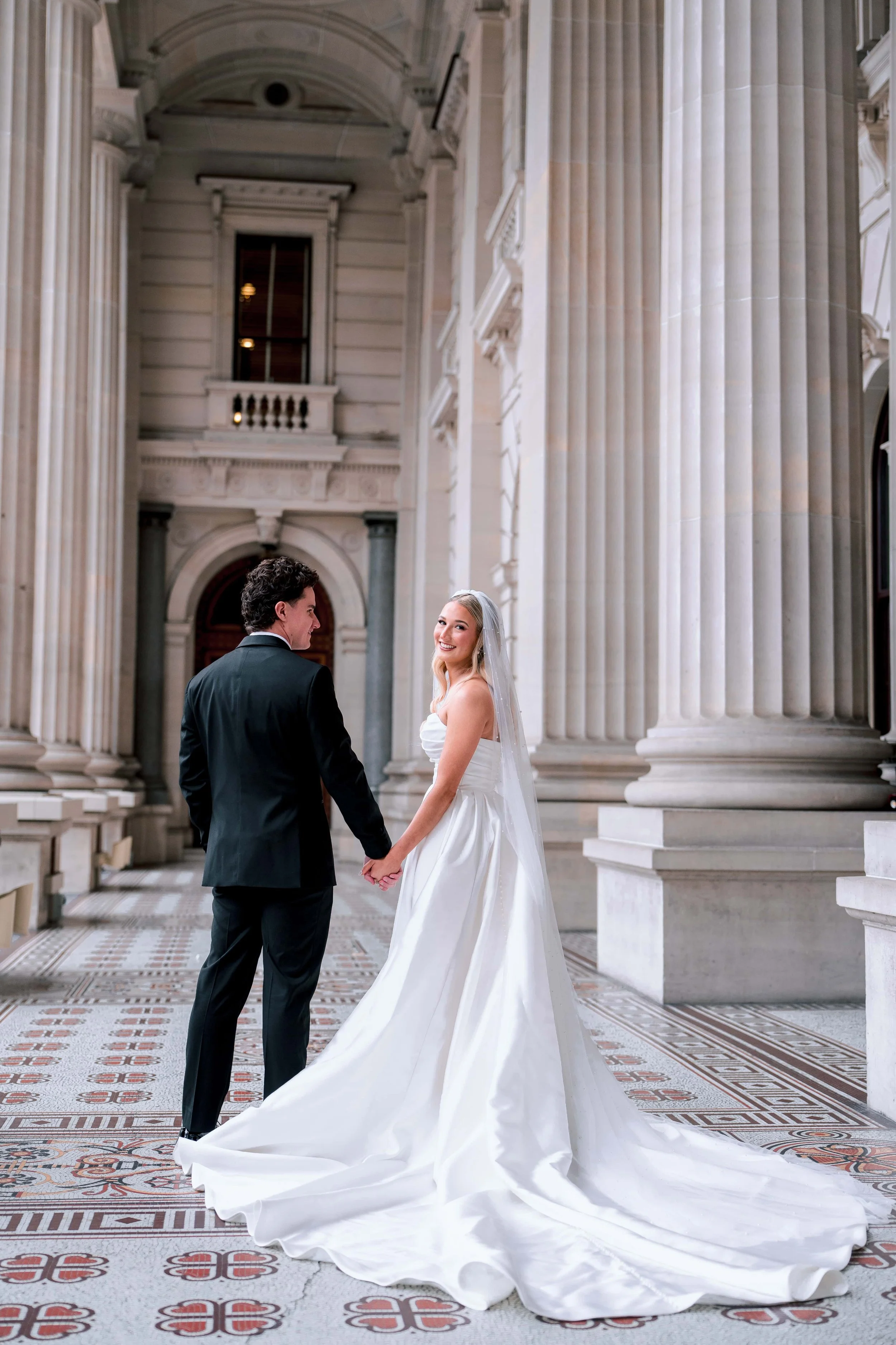 A bride and groom holding hands and looking at each other inside a grand building with large columns and intricate tile flooring.