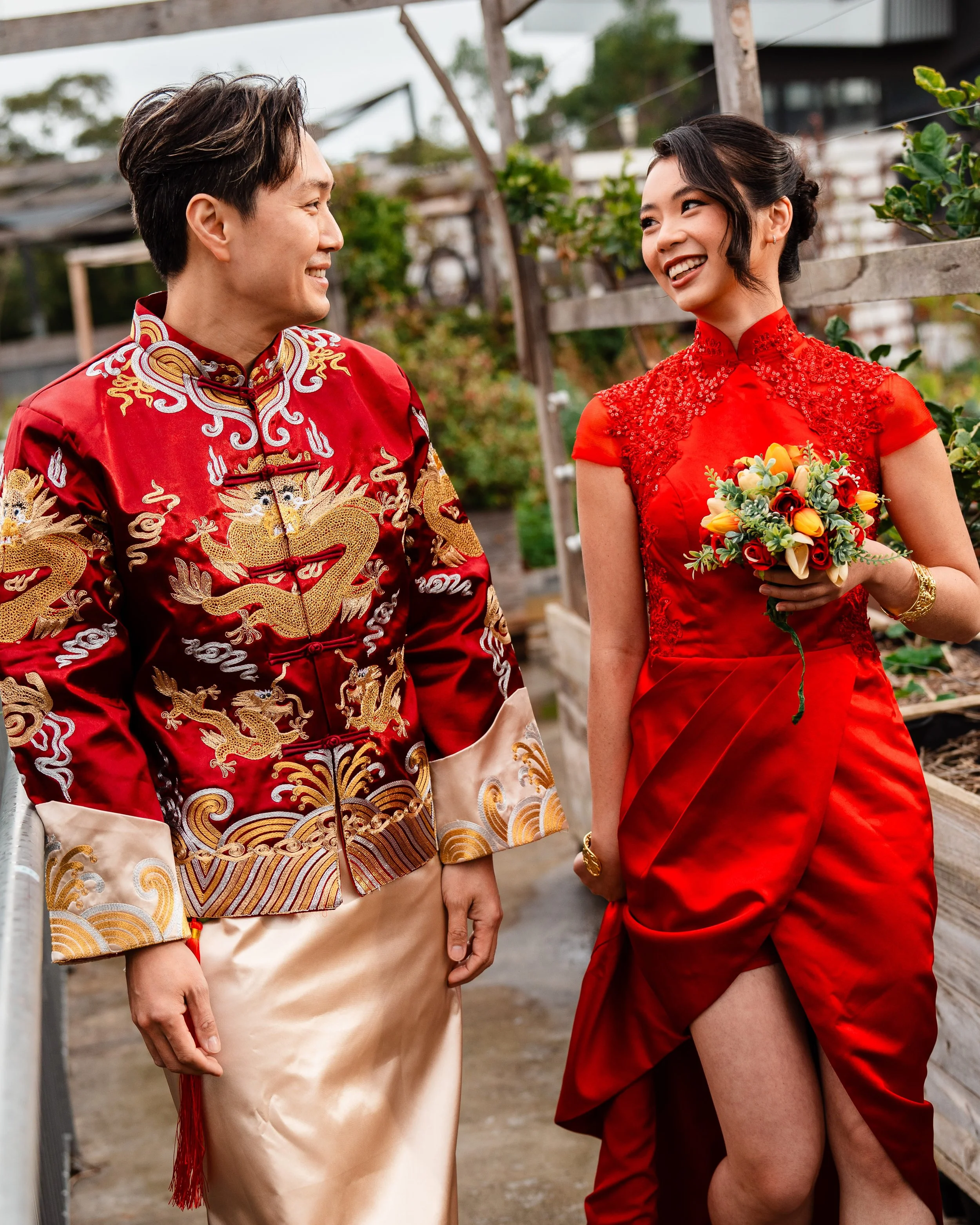 A man and a woman in traditional Chinese clothing standing outdoors and smiling at each other, with greenery and wooden structures in the background.