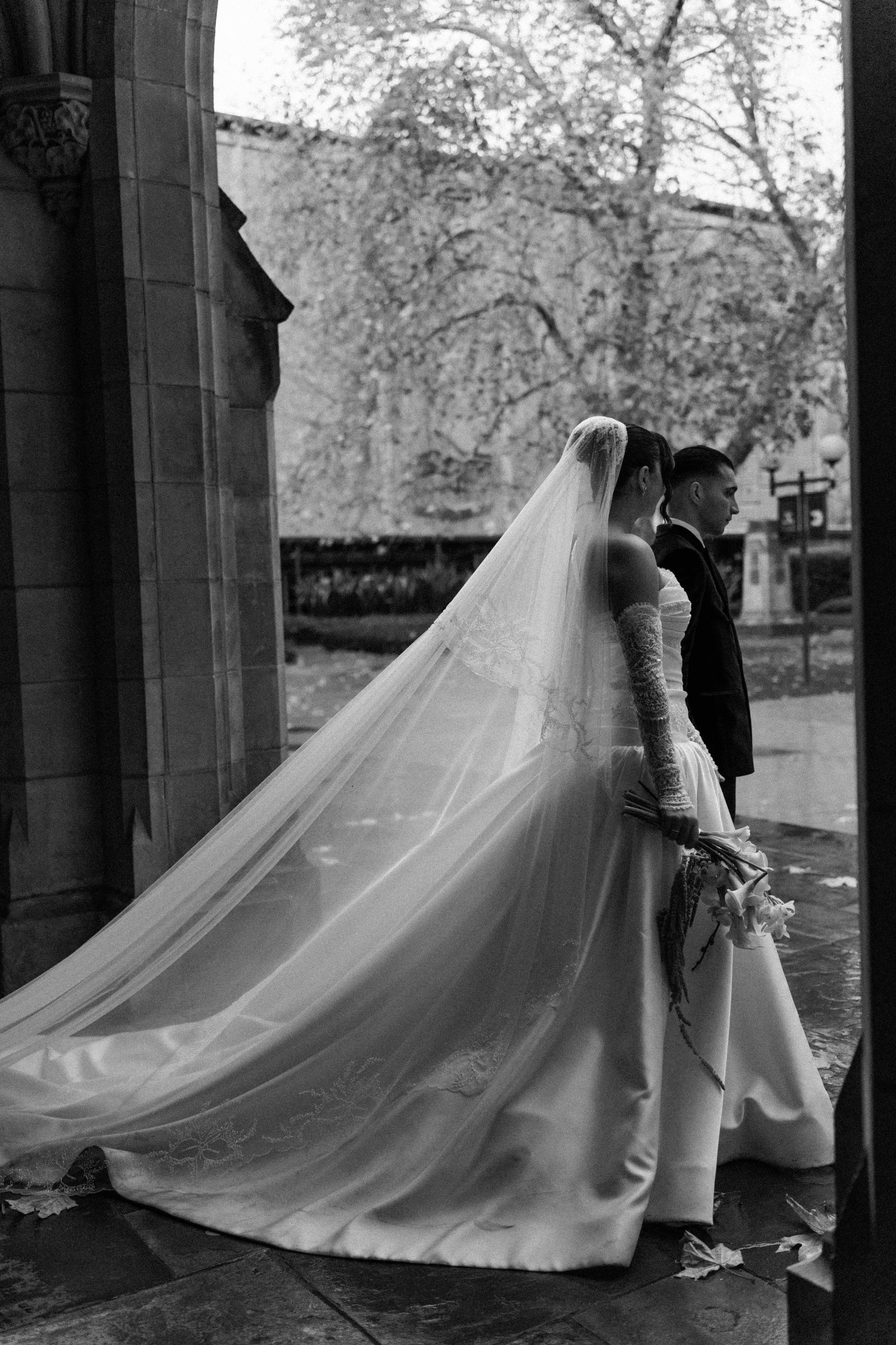 A black-and-white photo of a bride and groom kissing outdoors at the Fitzroy Gardens, Melbourne, the groom in a suit and the bride in a strapless wedding dress.