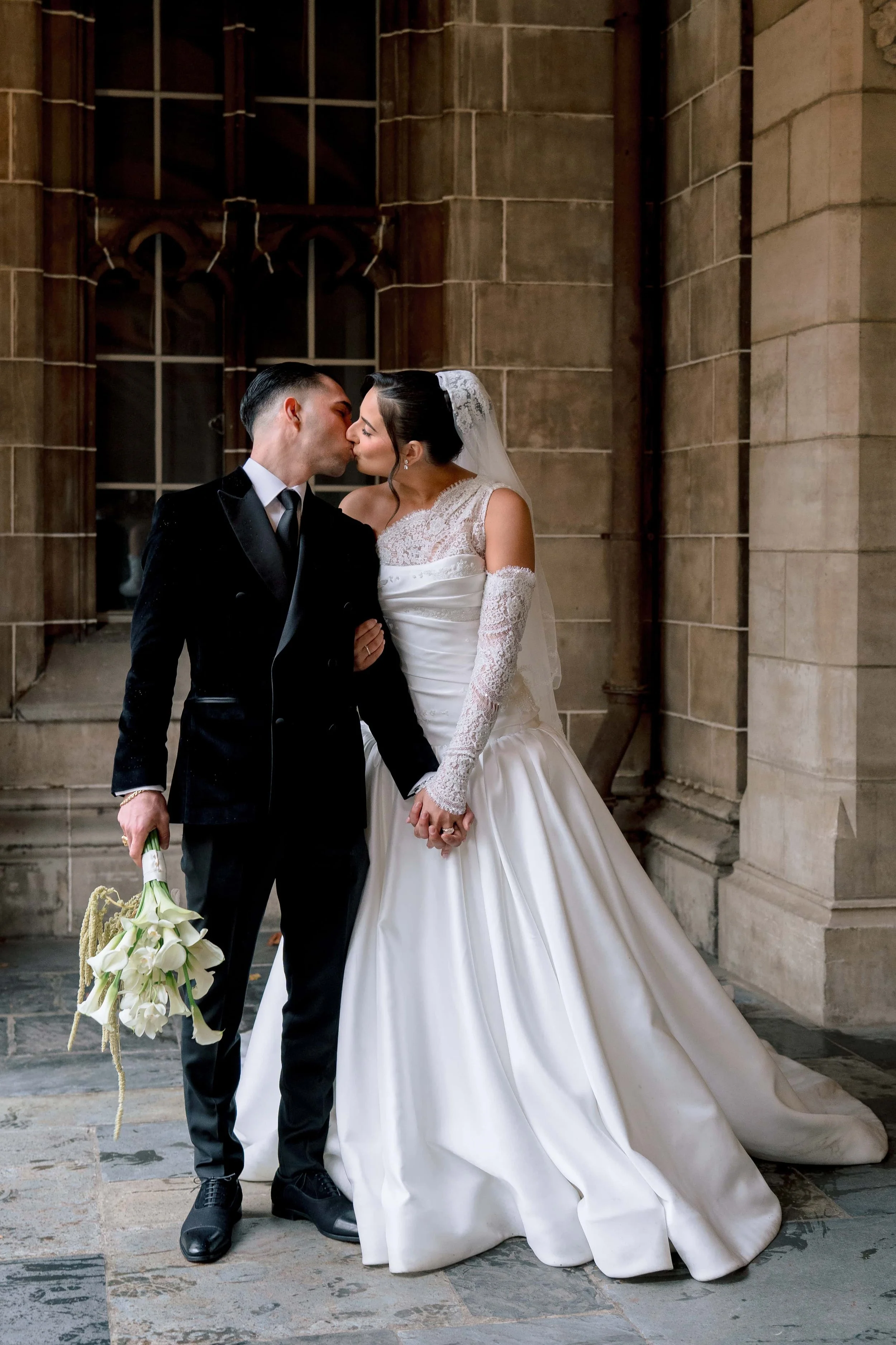 A bride and groom kissing outside a stone building. The bride is holding a bouquet of white calla lilies, wearing a white gown and veil. The groom is dressed in a black tuxedo.