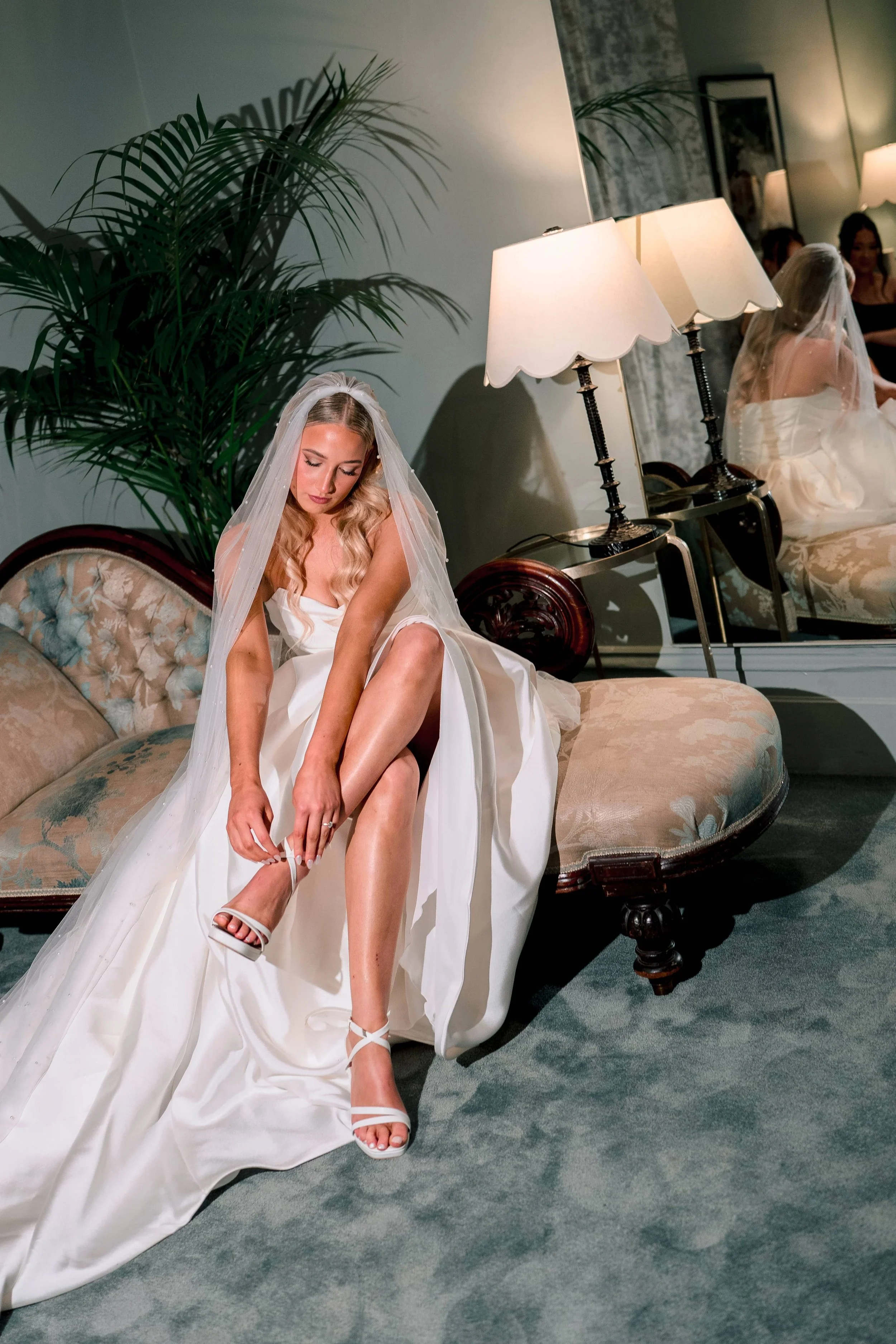 A bride in a white wedding dress sitting on an antique sofa, putting on her sandal with the other foot. She is wearing a veil and has long, wavy blonde hair. There is a large green plant behind her, and a mirror and lamps on a side table next to her. Several women are reflected in the mirror, sitting and chatting.