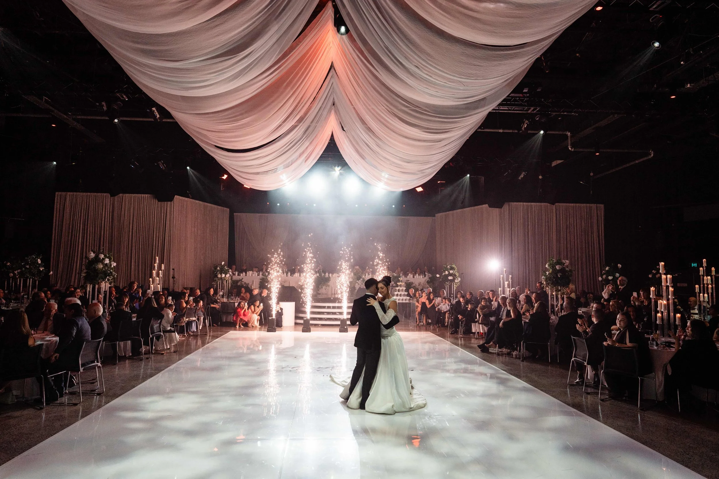 A bride and groom dancing in the center of a large, elegant banquet hall decorated with draped fabric ceiling, floral arrangements, and candlelit tables, with a packed audience watching.