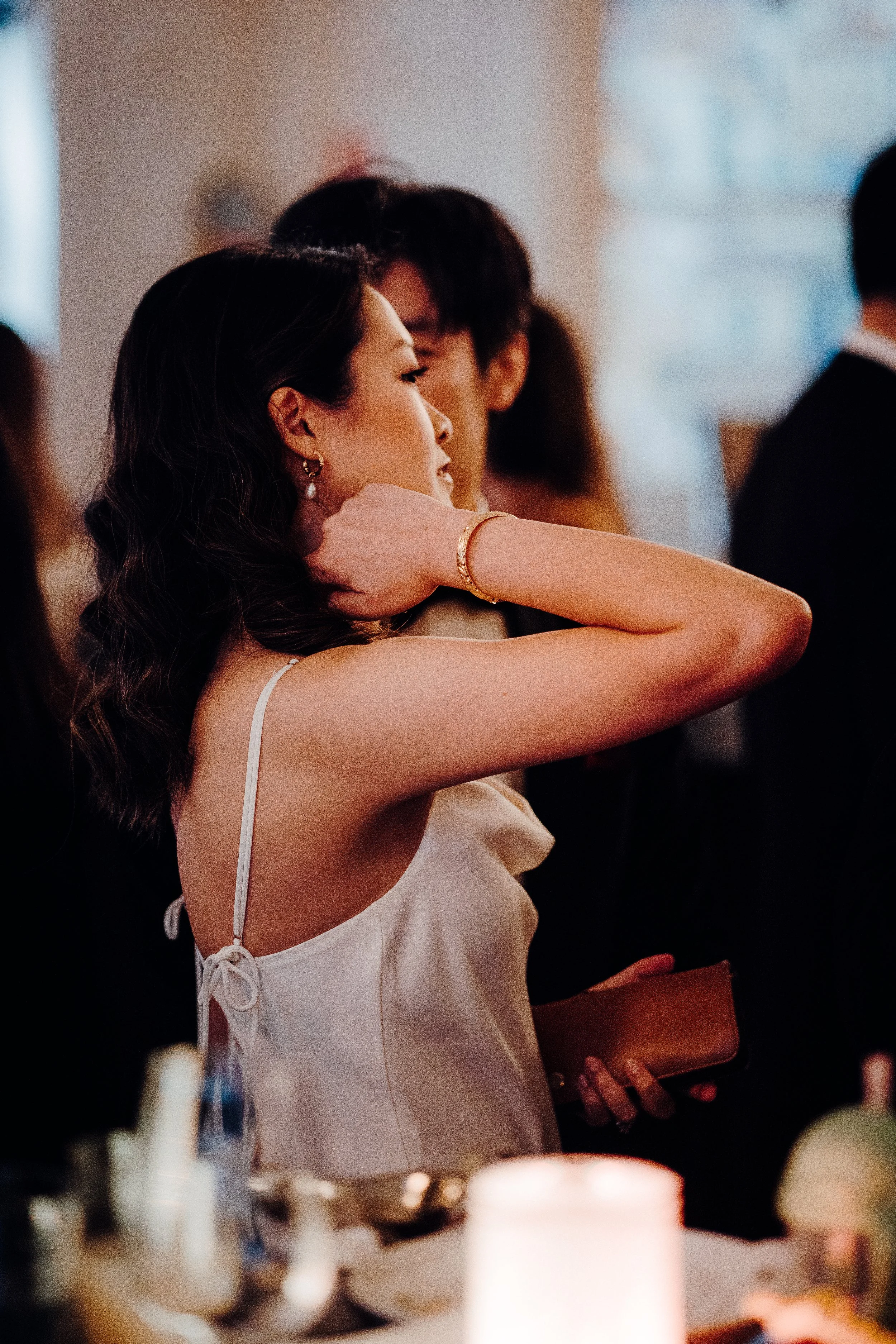 A woman with dark wavy hair and wearing earrings and a bracelet, dressed in a white dress with thin straps and a bow at the back, is adjusting her hair or neck while holding a purse in her left hand. She is standing indoors, surrounded by blurred figures in a social setting.