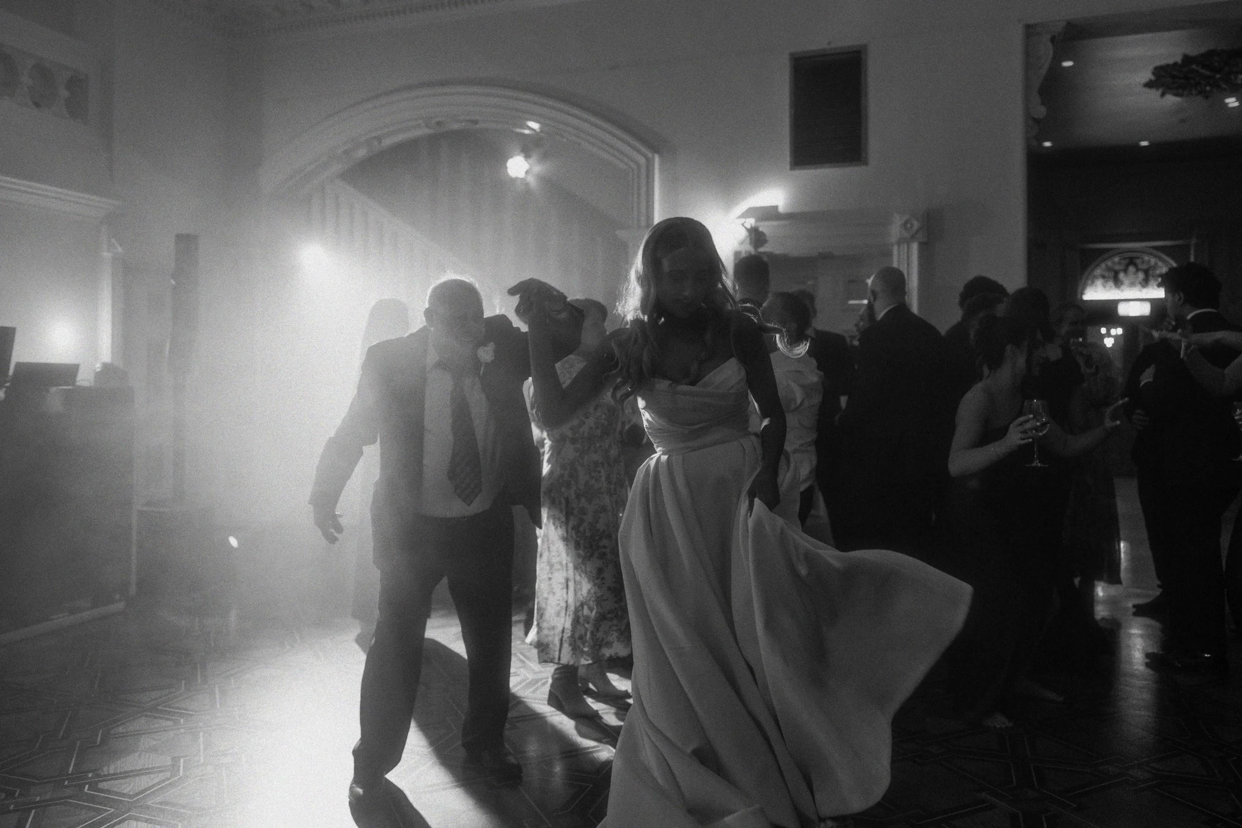 People dancing at a wedding reception in a dimly lit hall with a woman in a white dress in the foreground.