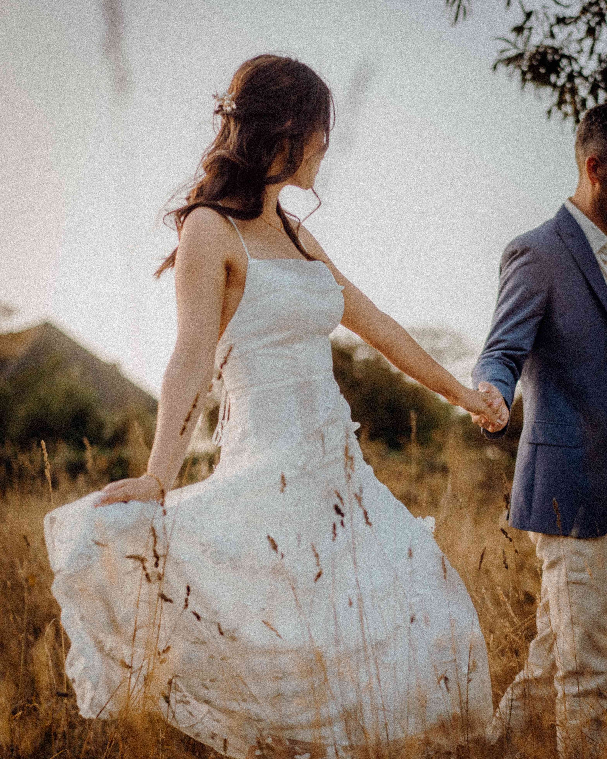 A woman in a white wedding dress holding hands with a man in a gray suit, walking in a field with tall grass during sunset.