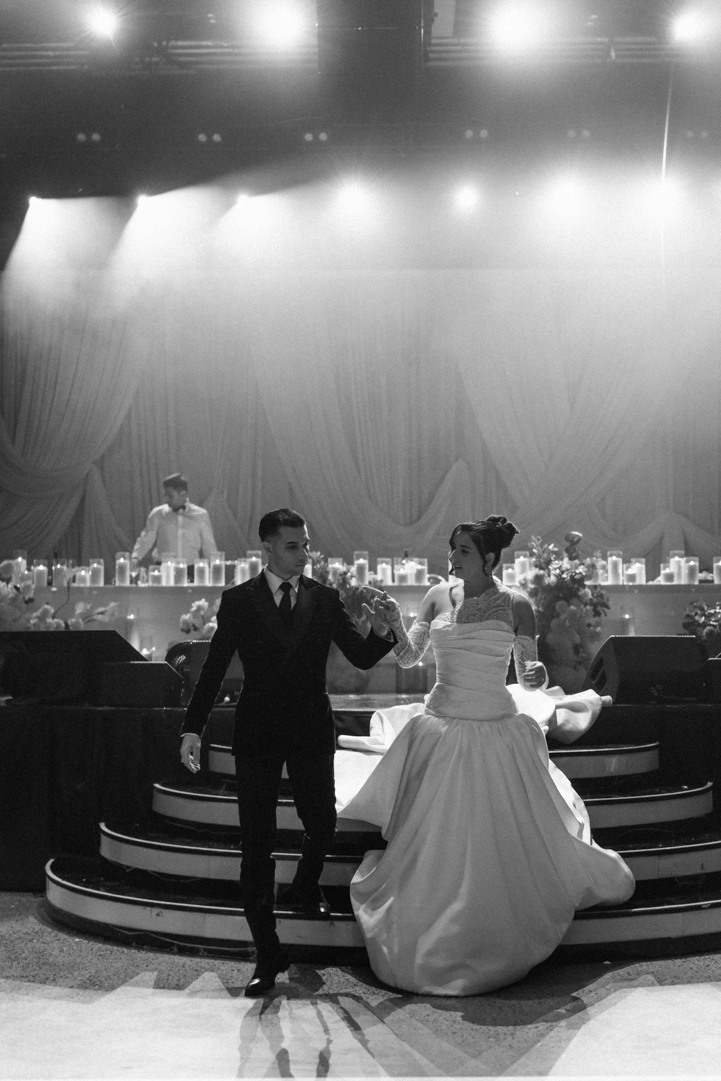 A black and white photo of a bride and groom dancing on stage during their wedding reception. The bride is in a strapless gown with a full skirt, and the groom is in a dark suit. They are holding hands and looking at each other, with a double staircase and a decorated background behind them. There are candles and floral arrangements on the stage.