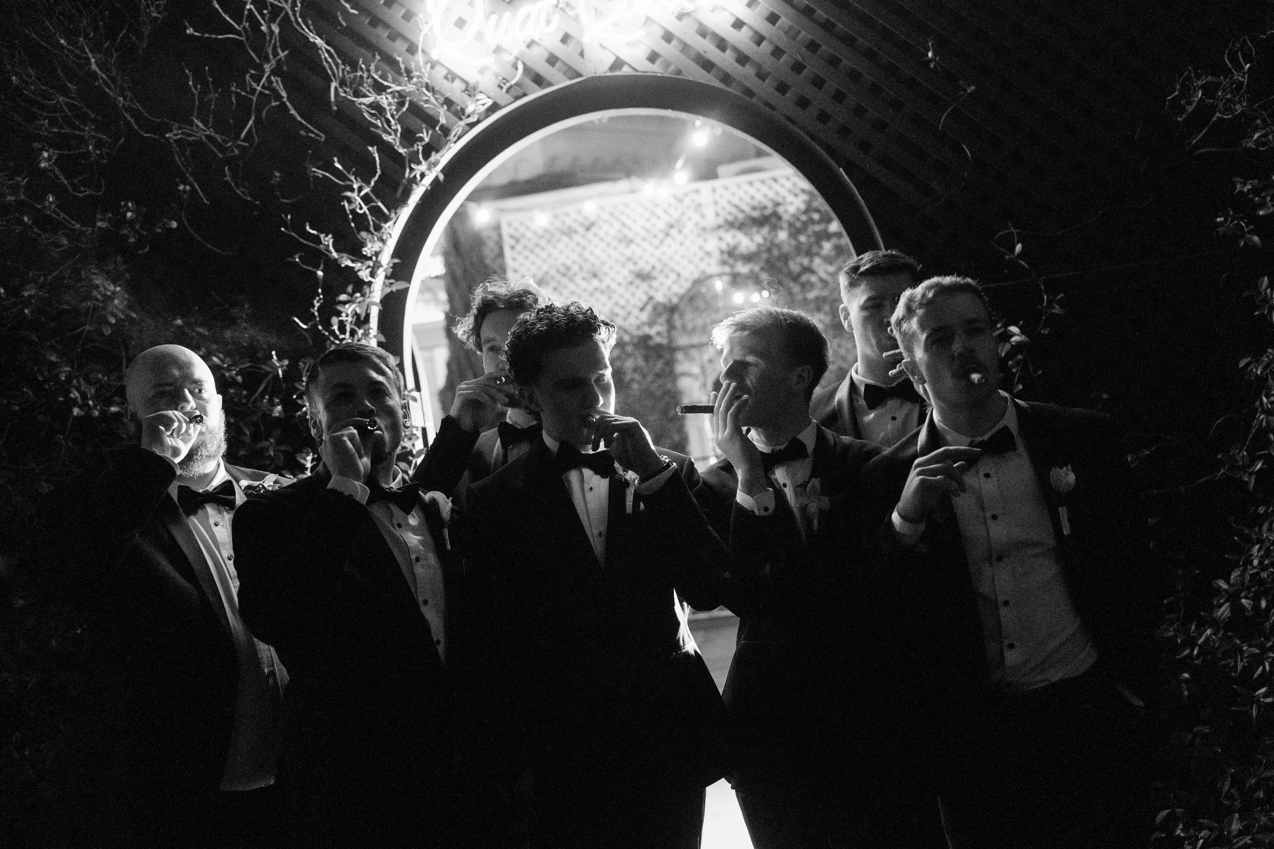Group of men dressed in tuxedos smoking cigars at a nighttime outdoor event, standing beneath an archway decorated with plants and lights.