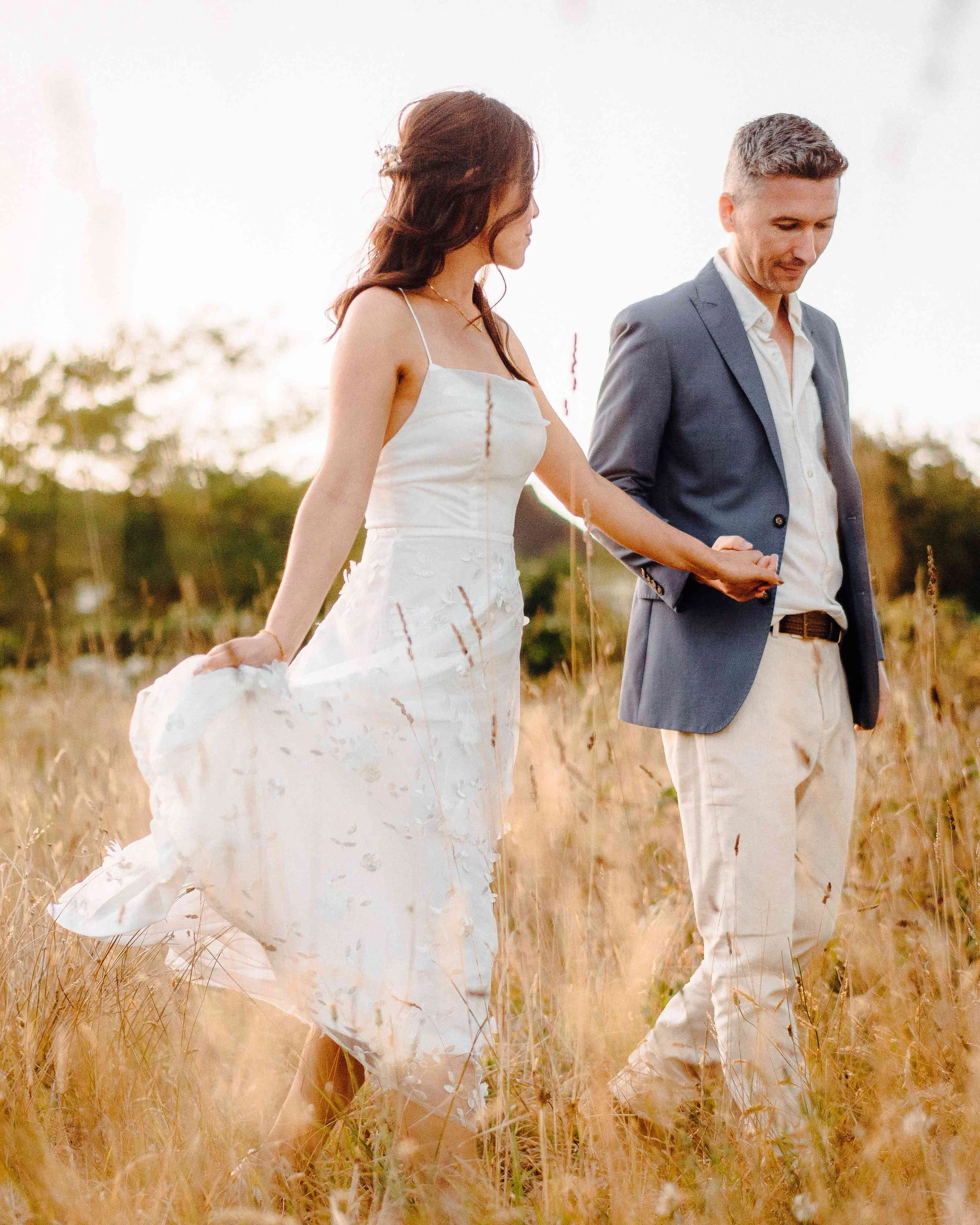 A couple holding hands in a field of tall, golden grass during sunset, with blurred trees in the background.