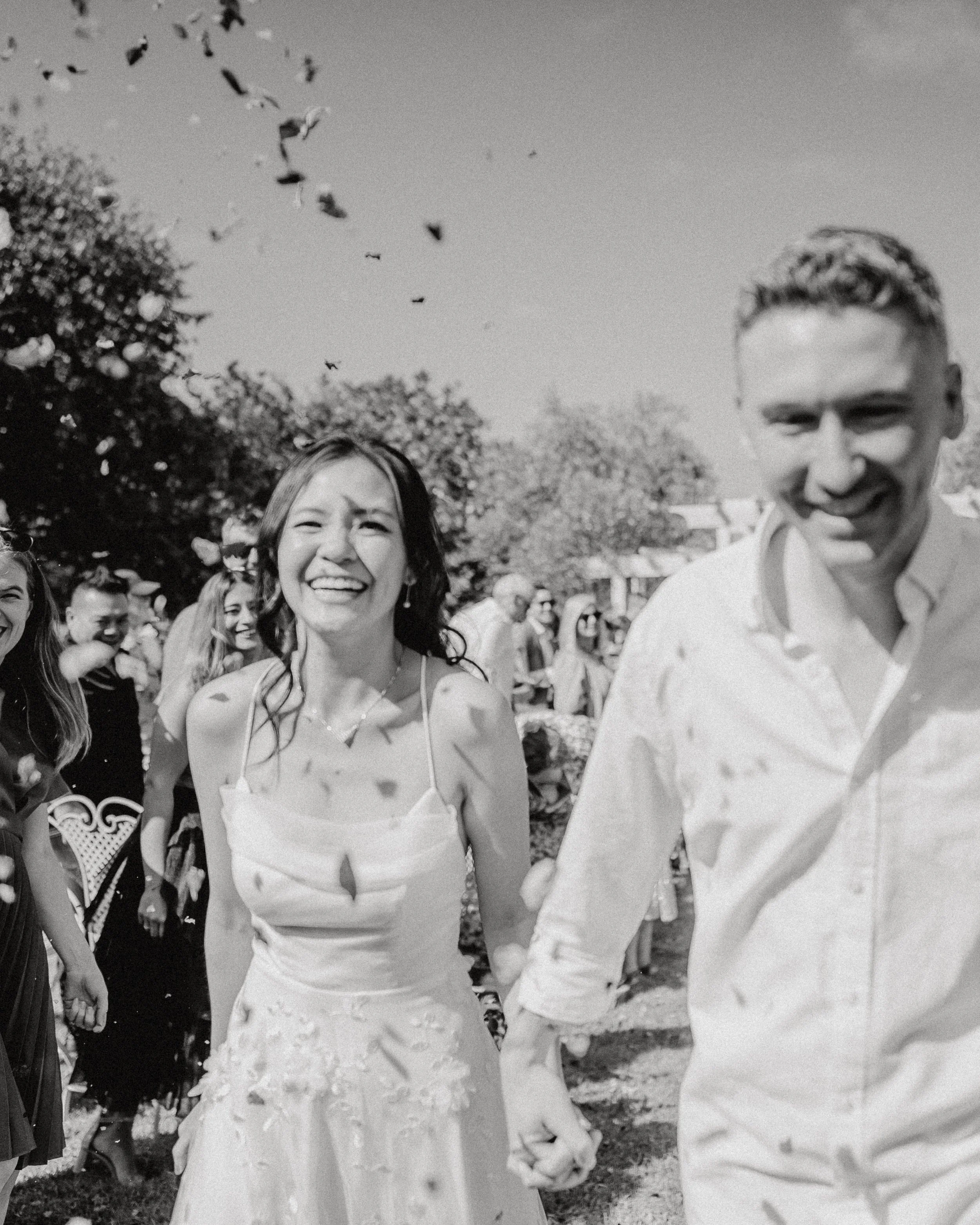 A black and white photo of a smiling bride and groom holding hands at their wedding, with guests in the background, outdoors on a sunny day.