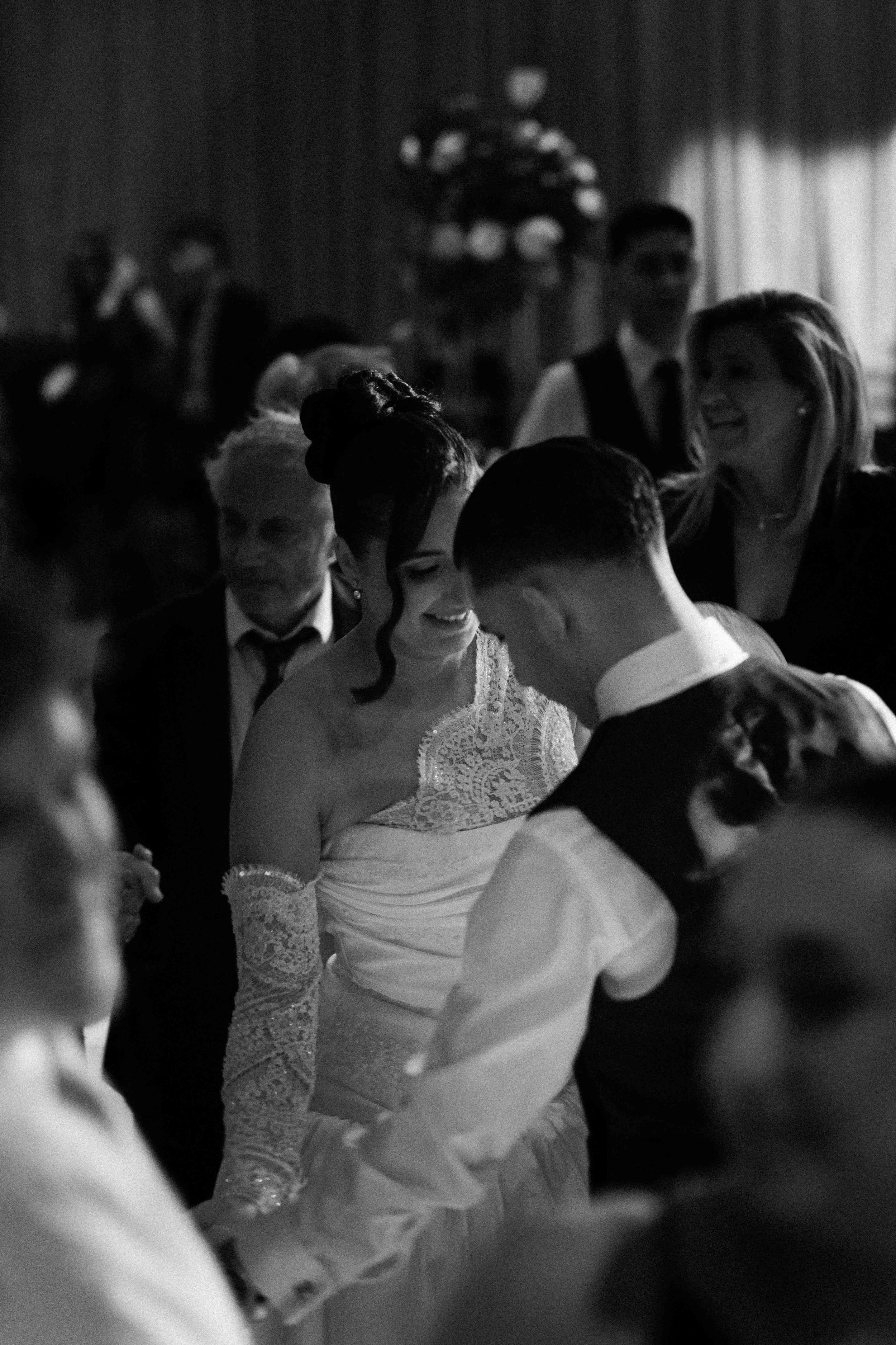 A black and white photo of a wedding reception with a bride and groom dancing, surrounded by guests in formal attire, some smiling and engaged in conversation.