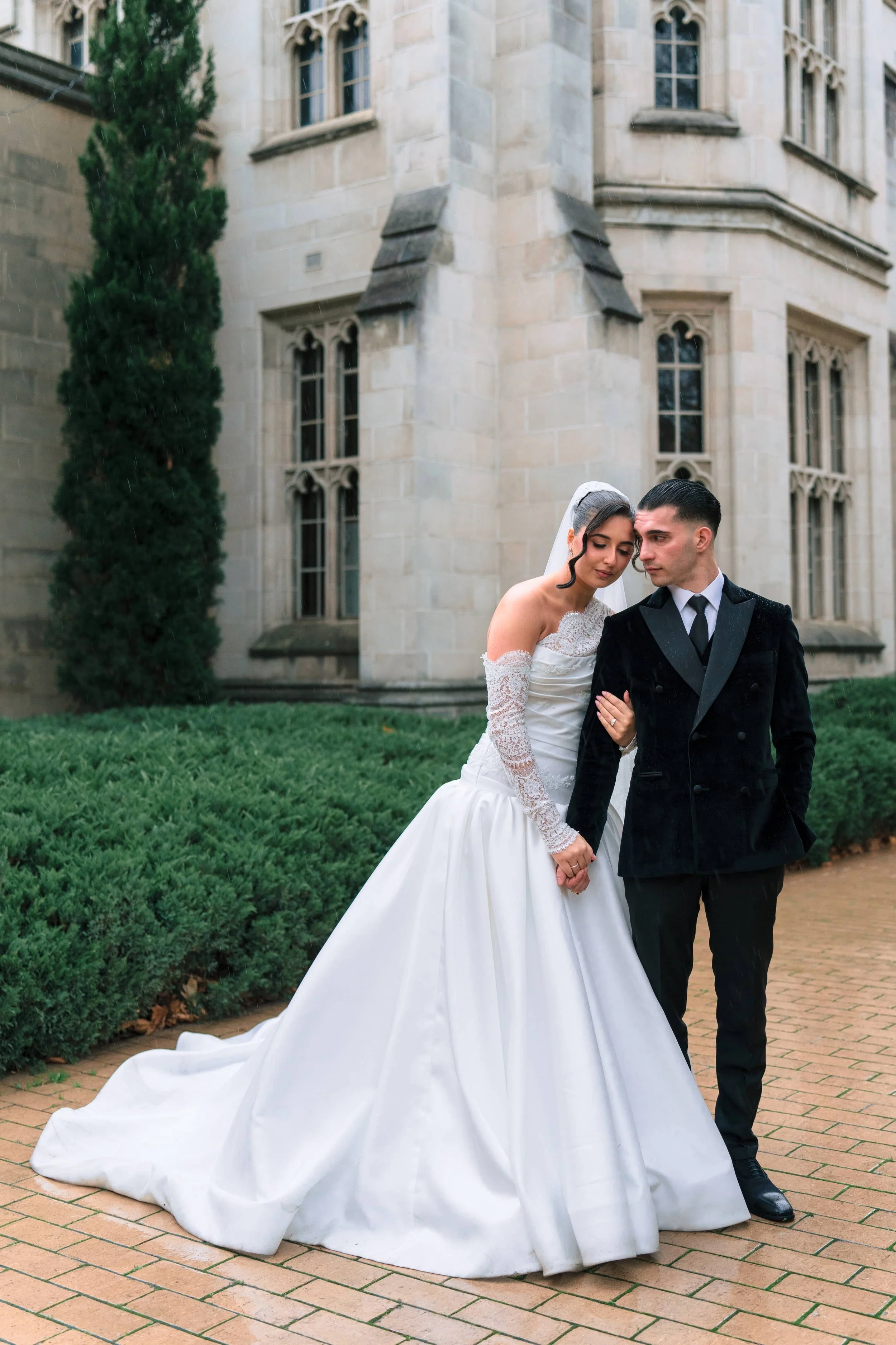 A bride and groom standing close together on a brick pathway outside a historic building with Gothic-style windows, with the bride wearing a white wedding dress and veil, and the groom in a black tuxedo.