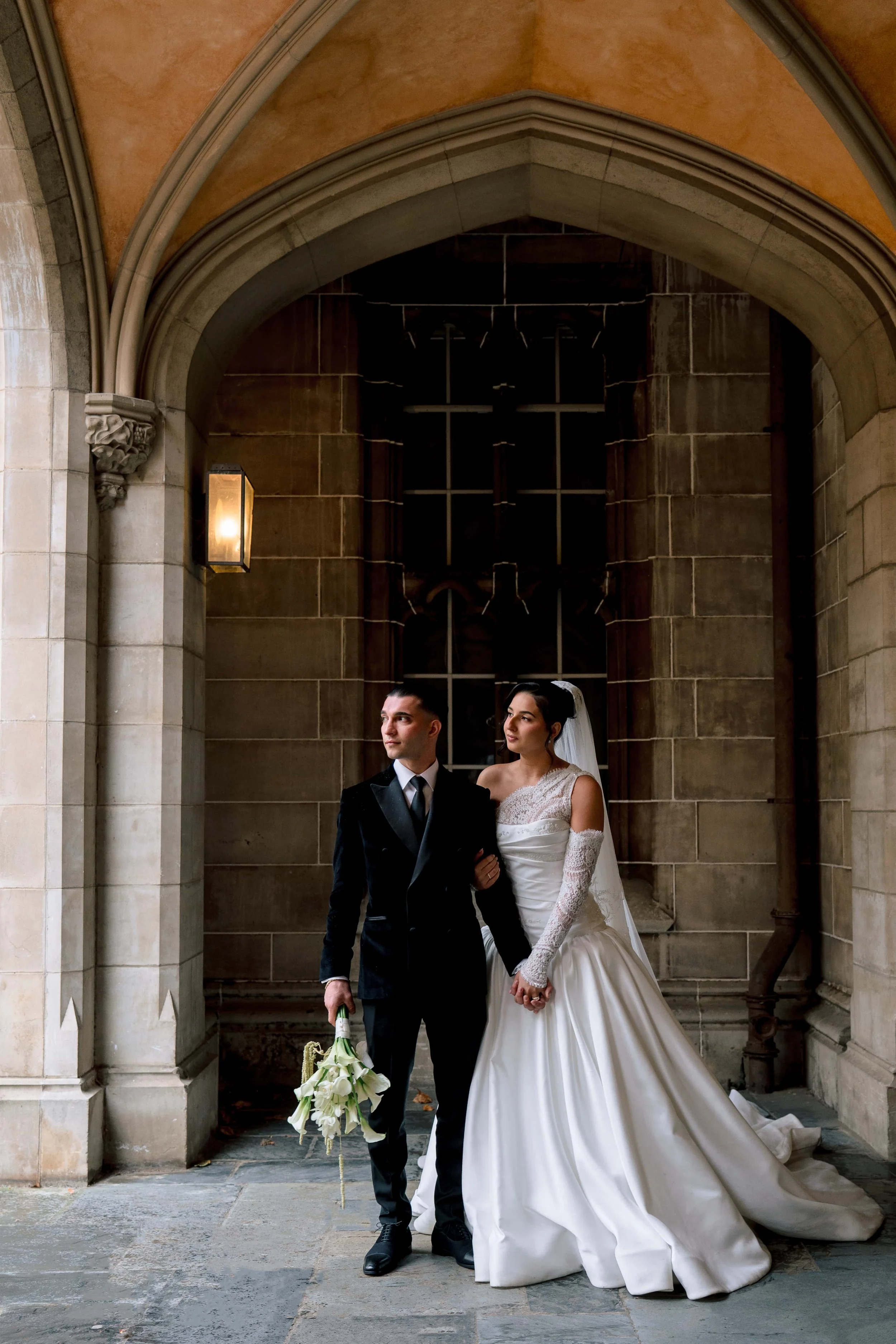 A bride and groom standing together in front of a church doorway, holding hands. The bride is in a white wedding dress with lace details and a veil, and the groom is in a black tuxedo. The groom is holding a bouquet of white calla lilies.