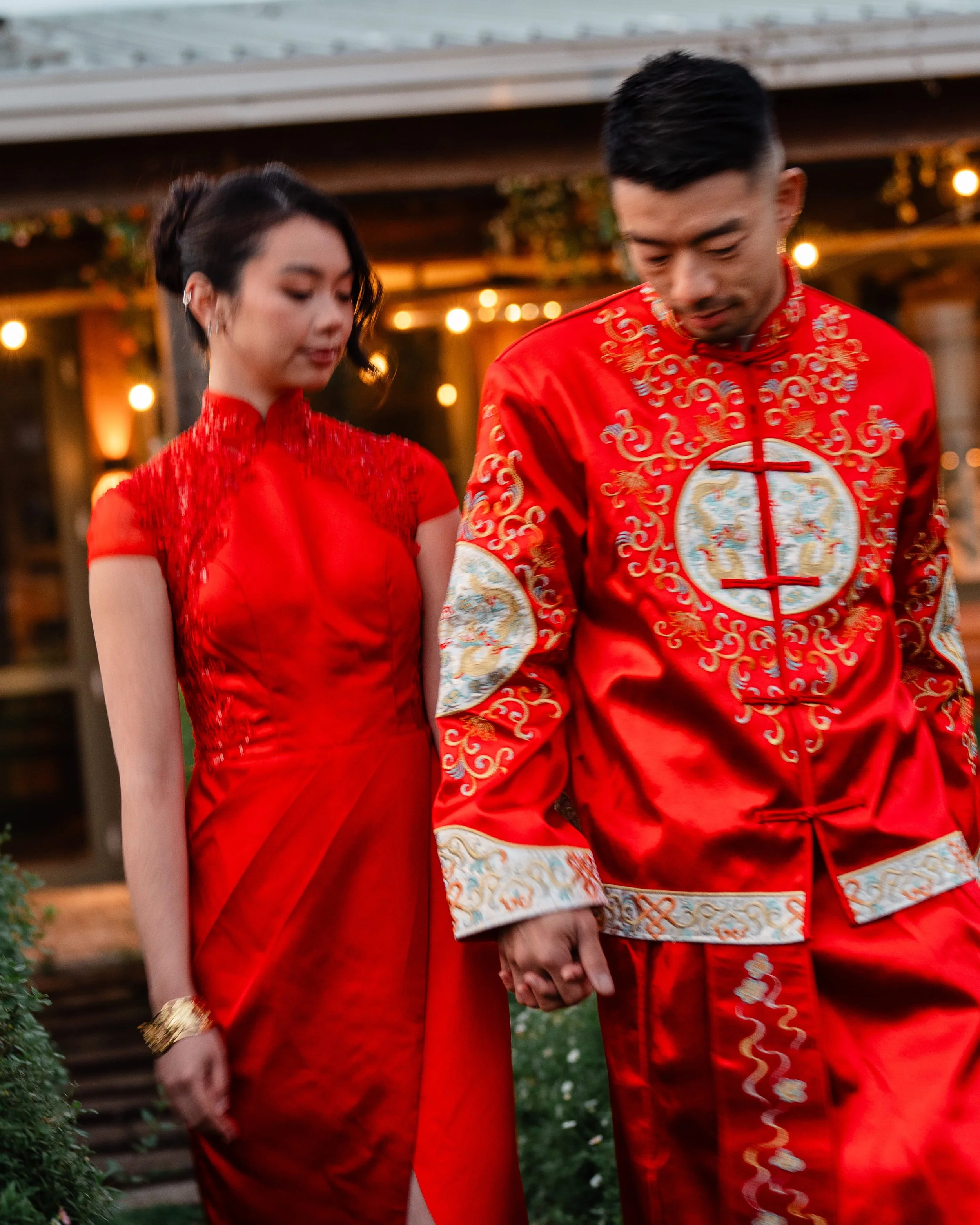 A couple dressed in traditional Chinese wedding attire, holding hands, outdoors in a garden setting, during evening with warm lights in the background.