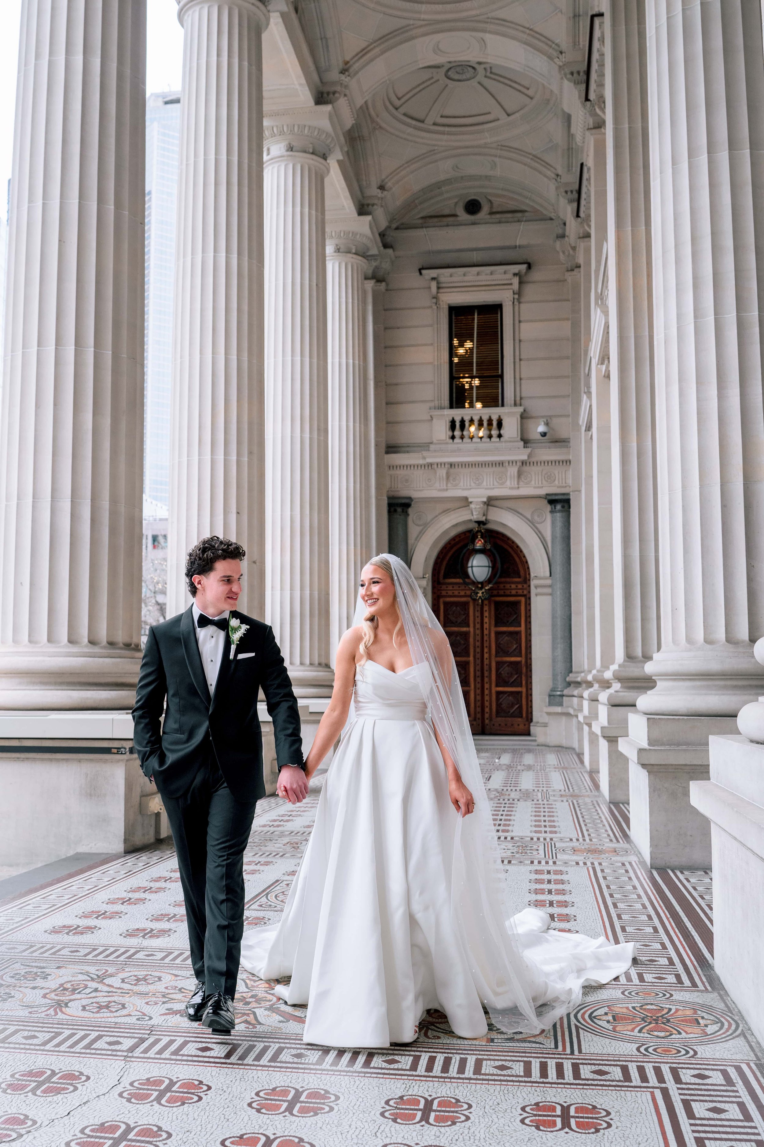 A bride and groom holding hands and walking together in front of a building with large columns, arches, and ornate tile floor. The groom is in a black tuxedo with a bow tie, and the bride is in a white wedding gown with a veil.