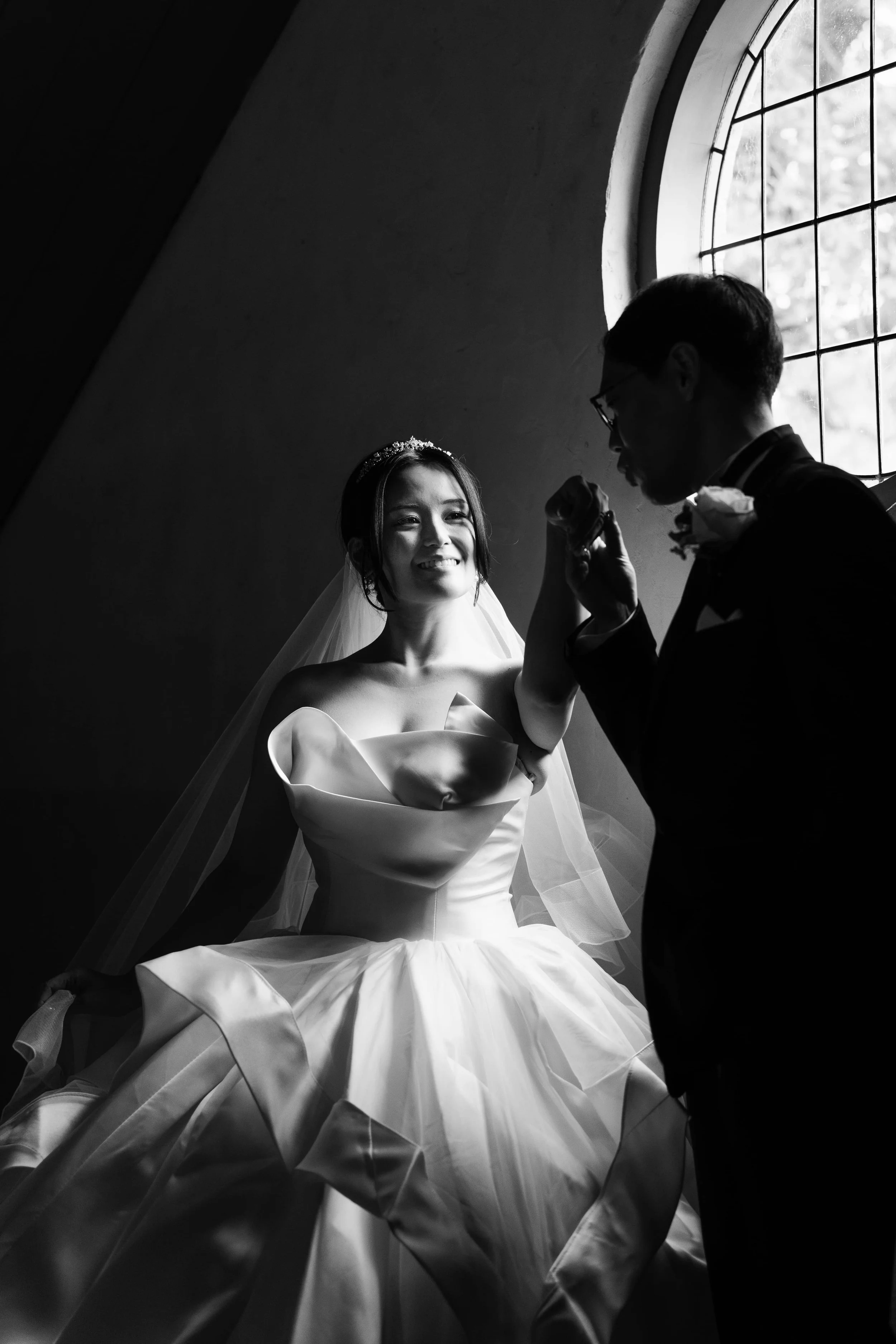 A black and white photo of a bride in a wedding dress with a veil and tiara, smiling, holding a bouquet, with a groom in a suit and glasses, near a large arched window with grid panes, inside a building with a curved wall.