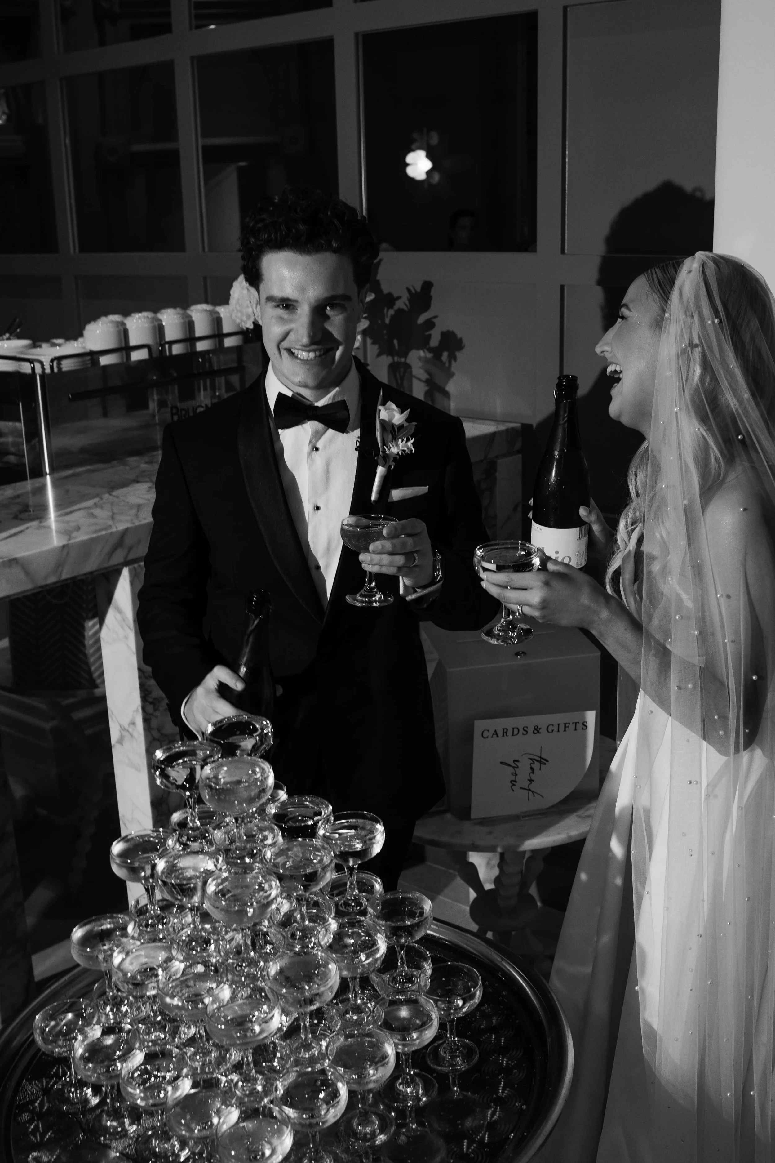 Black and white photo of a man and woman in formal attire at a celebration. The man in a tuxedo and bow tie is holding a glass of drink and smiling. The woman, in a wedding dress and veil, is laughing and holding a bottle and a glass. They are standi