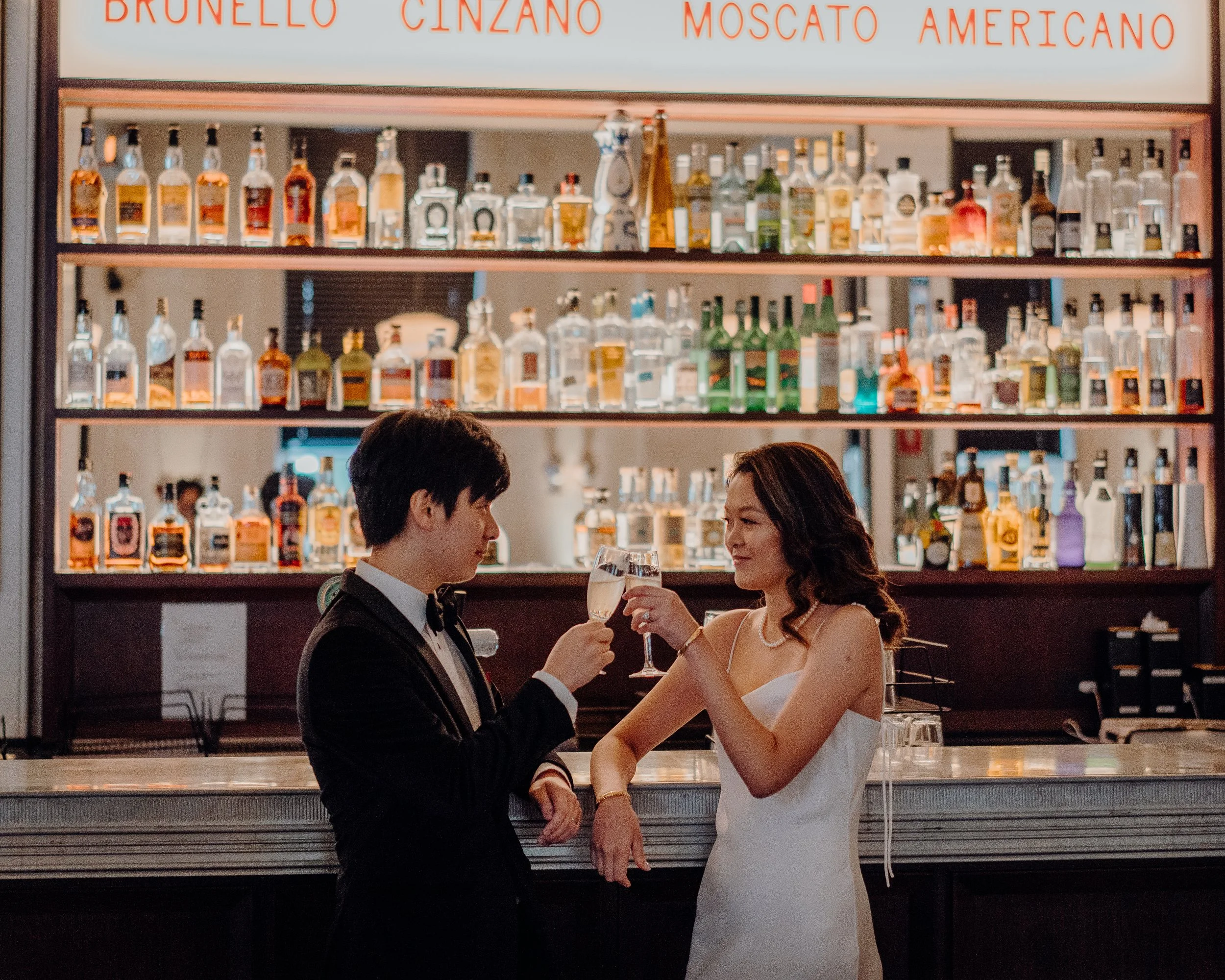 A man in a tuxedo and a woman in a white dress are raising glasses of wine in a toast at a formal event in a restaurant or bar. They are seated at a counter with a large wall of liquor bottles behind them, illuminated and decorated with a large sign 