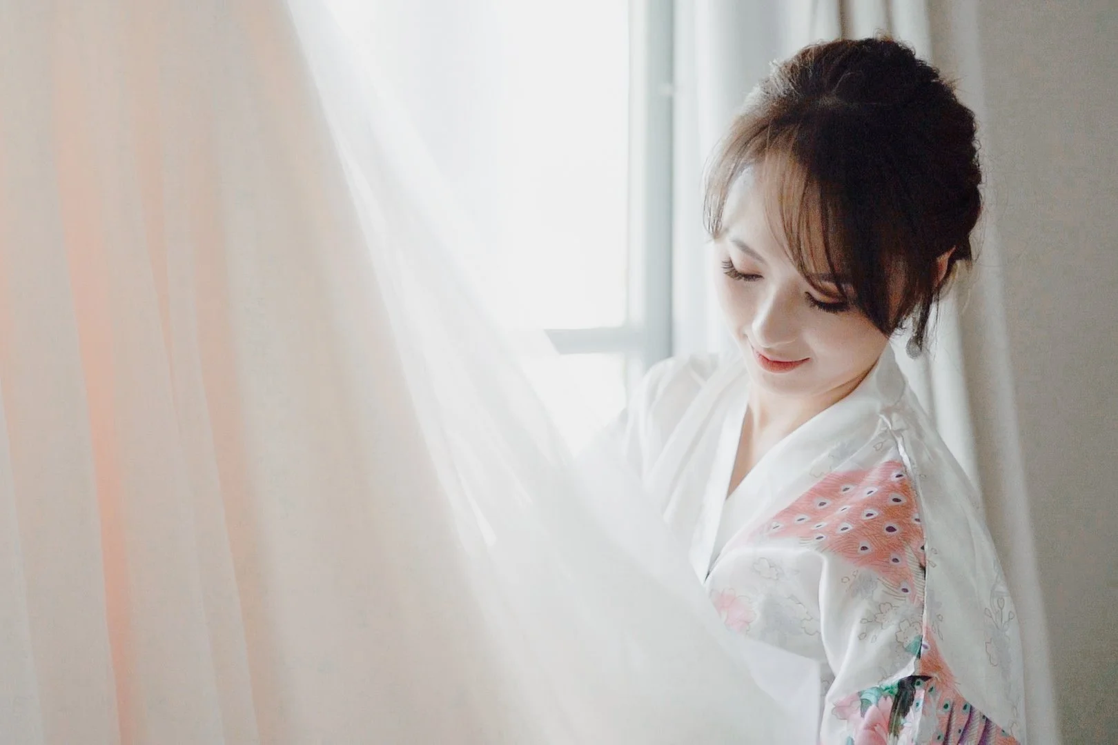 A woman with short dark hair looking down and smiling, wearing a white kimono with pastel-colored peacock feather pattern.
