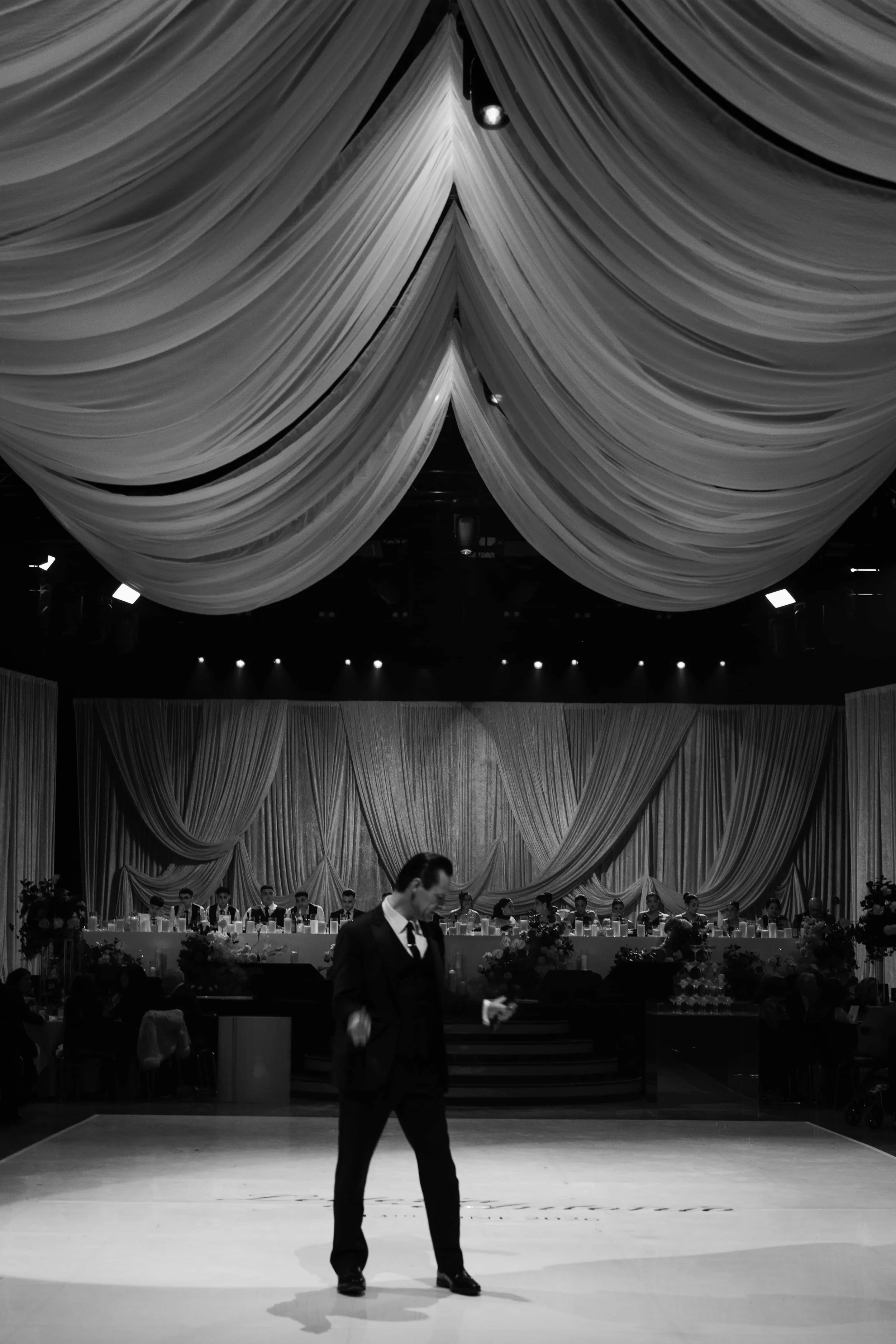 Black and white photo of a formal event with a decorated stage featuring draped curtains and a long table with seated individuals. A man dressed in a tuxedo is walking in the foreground.