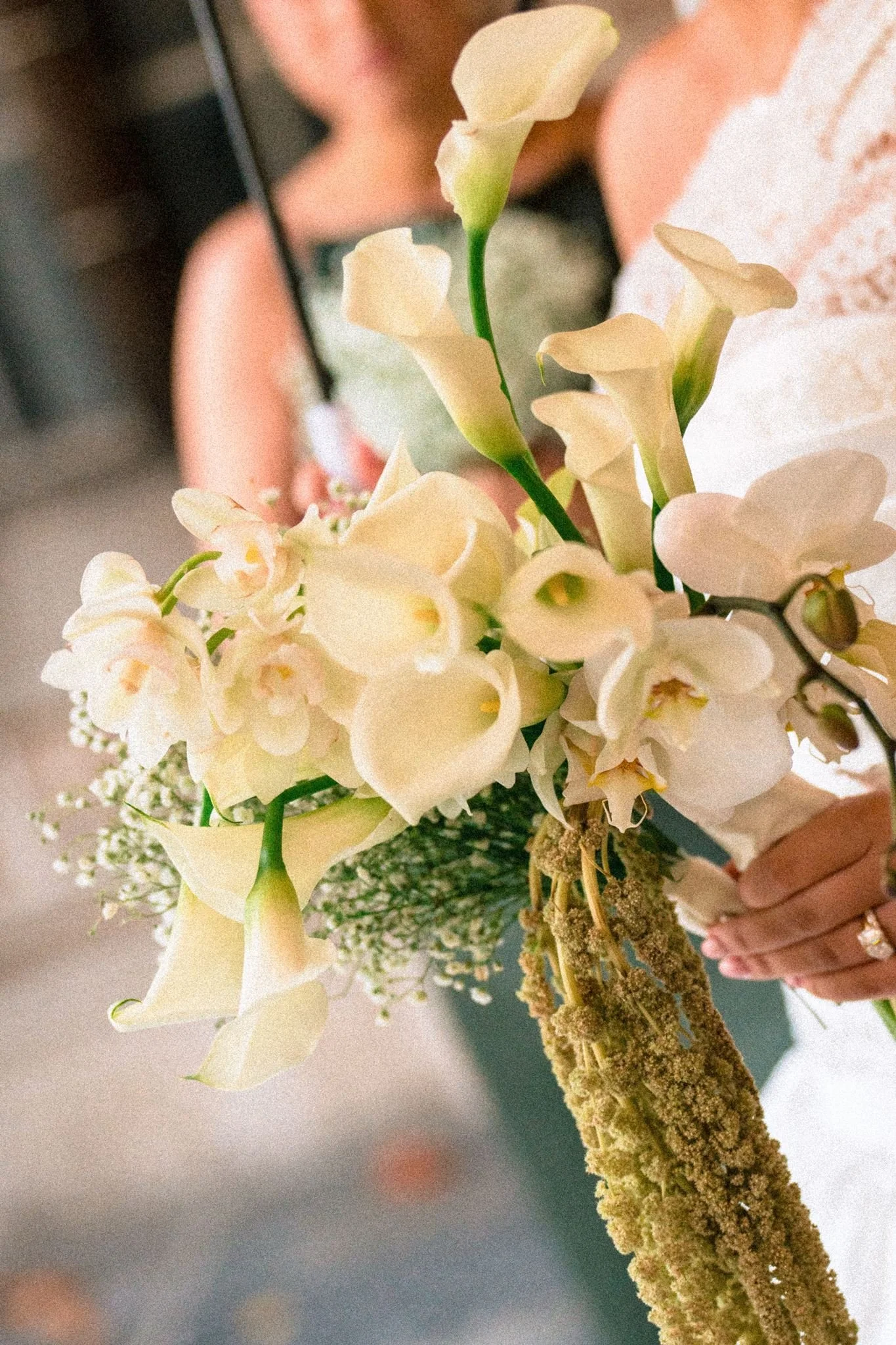 A close-up of white orchids and a mossy branch in the background, held by a person wearing a wedding ring and a lace dress.