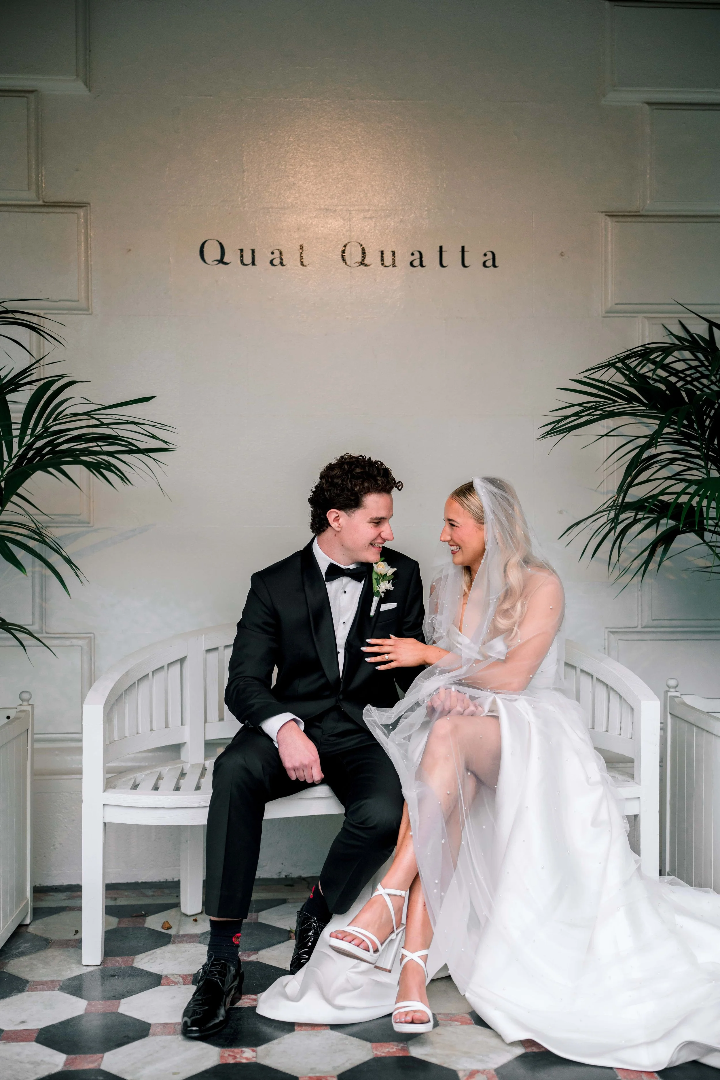 A wedding couple sitting on a white bench, smiling at each other. The groom wears a black tuxedo and the bride wears a white wedding gown and veil, with her leg raised showing white high-heeled shoes. There are plants on each side and a sign behind t
