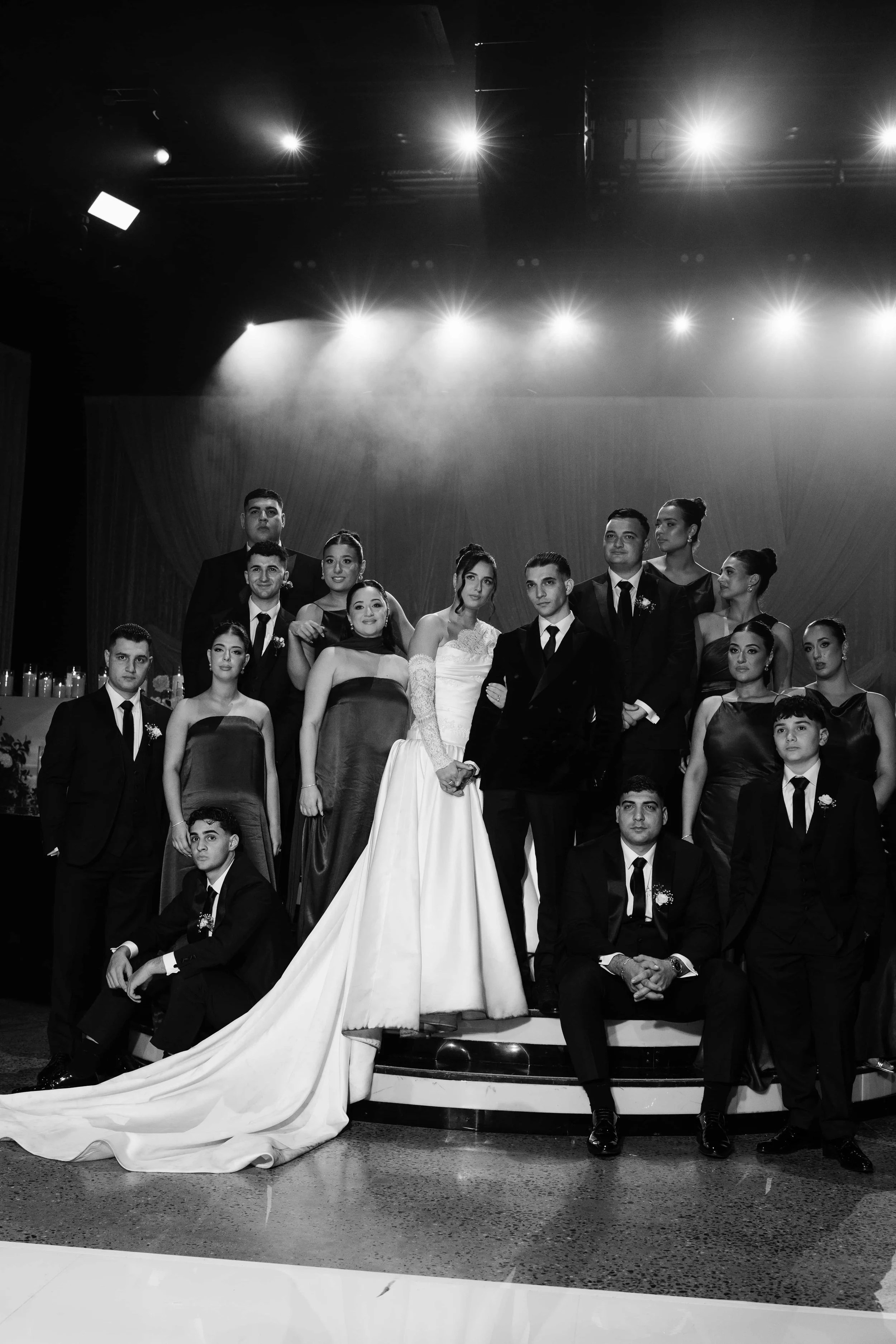 A group of formally dressed people, including a bride in a wedding gown and groom in a suit, posing on stage for a wedding photo with spotlights overhead, in black and white.