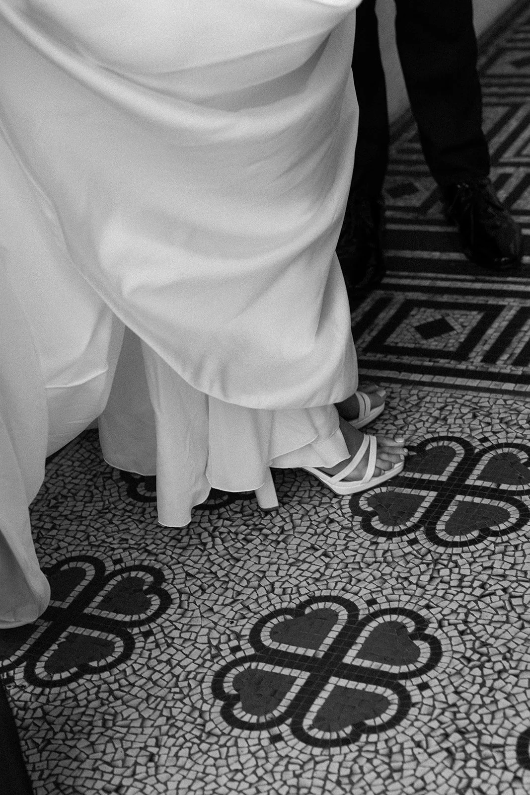 Close-up of the lower part of a wedding dress, a woman's foot in a high-heeled sandal, and patterned floor tiles.