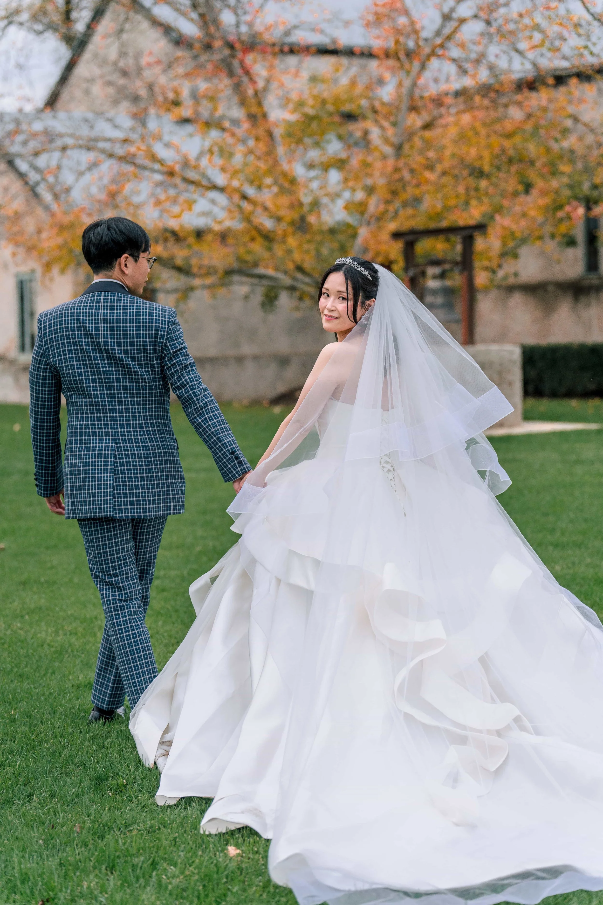 A bride in a white wedding gown with a long, flowing skirt and a veil, holding hands with a groom in a blue checkered suit, walking outdoors on green grass with autumn trees in the background.