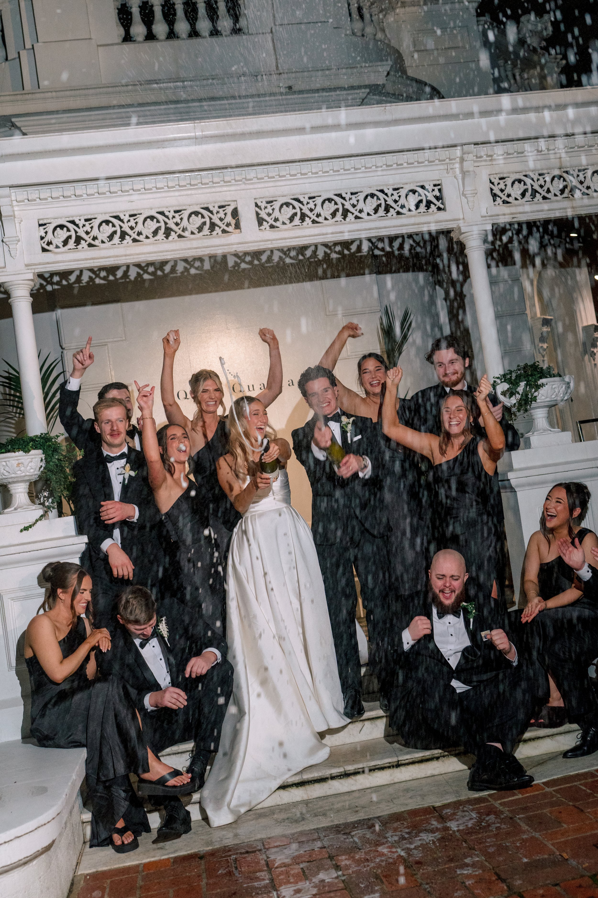 People celebrating on the staircase at a wedding, some are dancing and holding champagne glasses, under a white gazebo with intricate woodwork, in rainy weather.