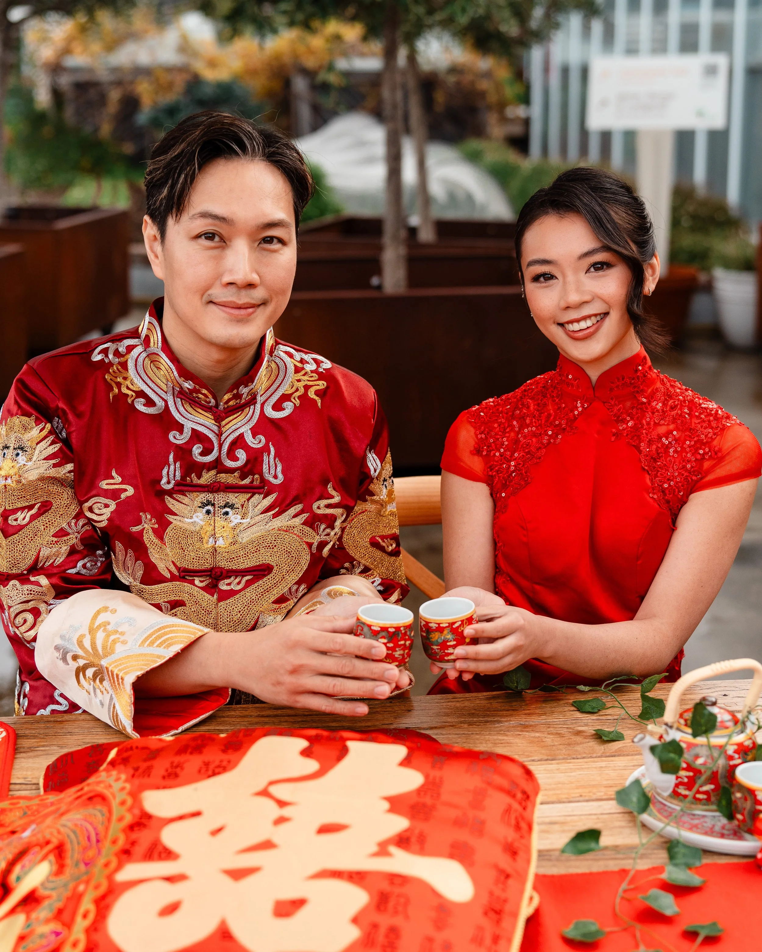 A man and woman dressed in traditional Chinese wedding attire, smiling and holding small cups, seated at a wooden table decorated with red and gold items, outdoors with autumn foliage in the background.
