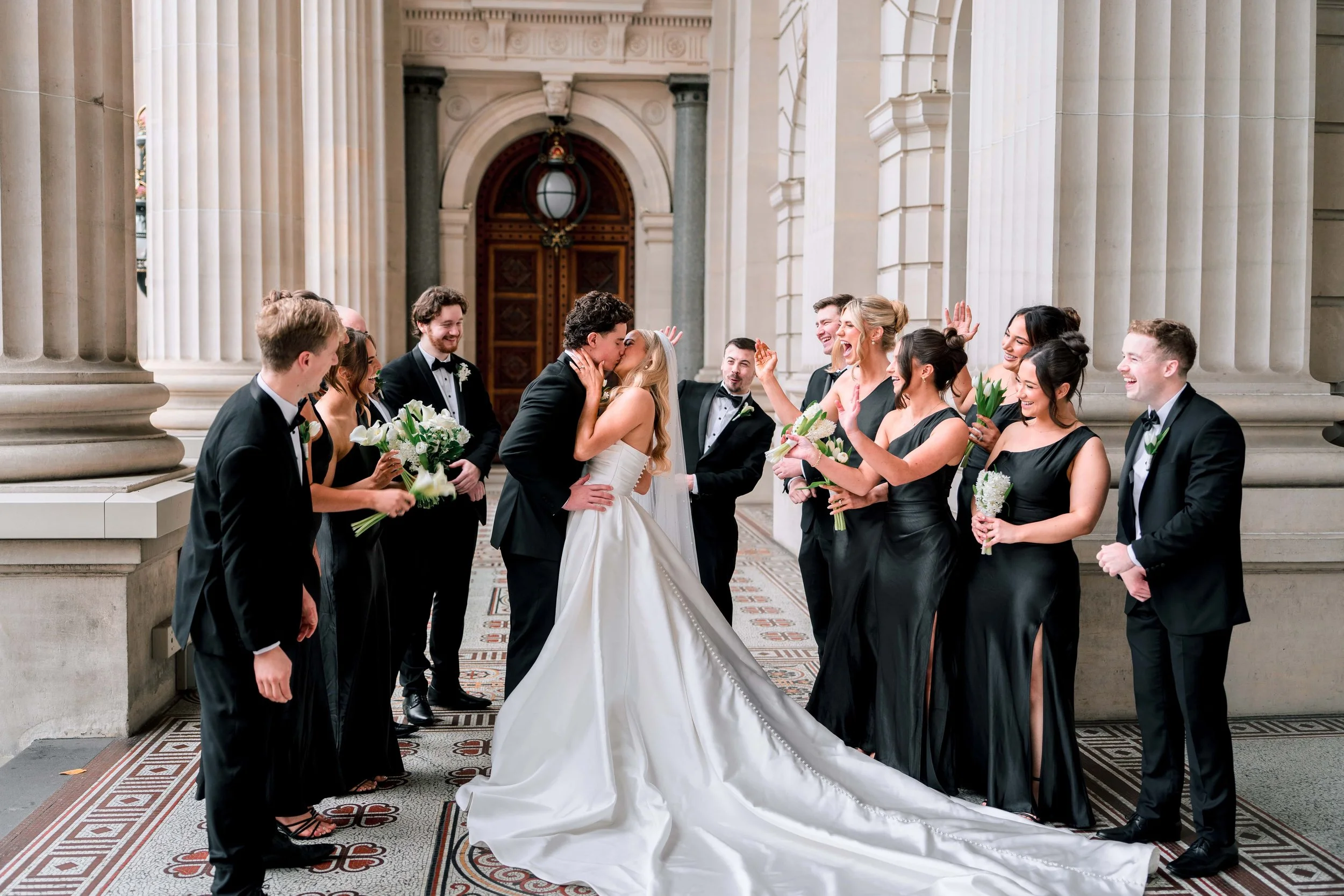 A wedding ceremony with a bride and groom kissing surrounded by bridesmaids and groomsmen, inside a grand building with large columns and ornate architectural details.