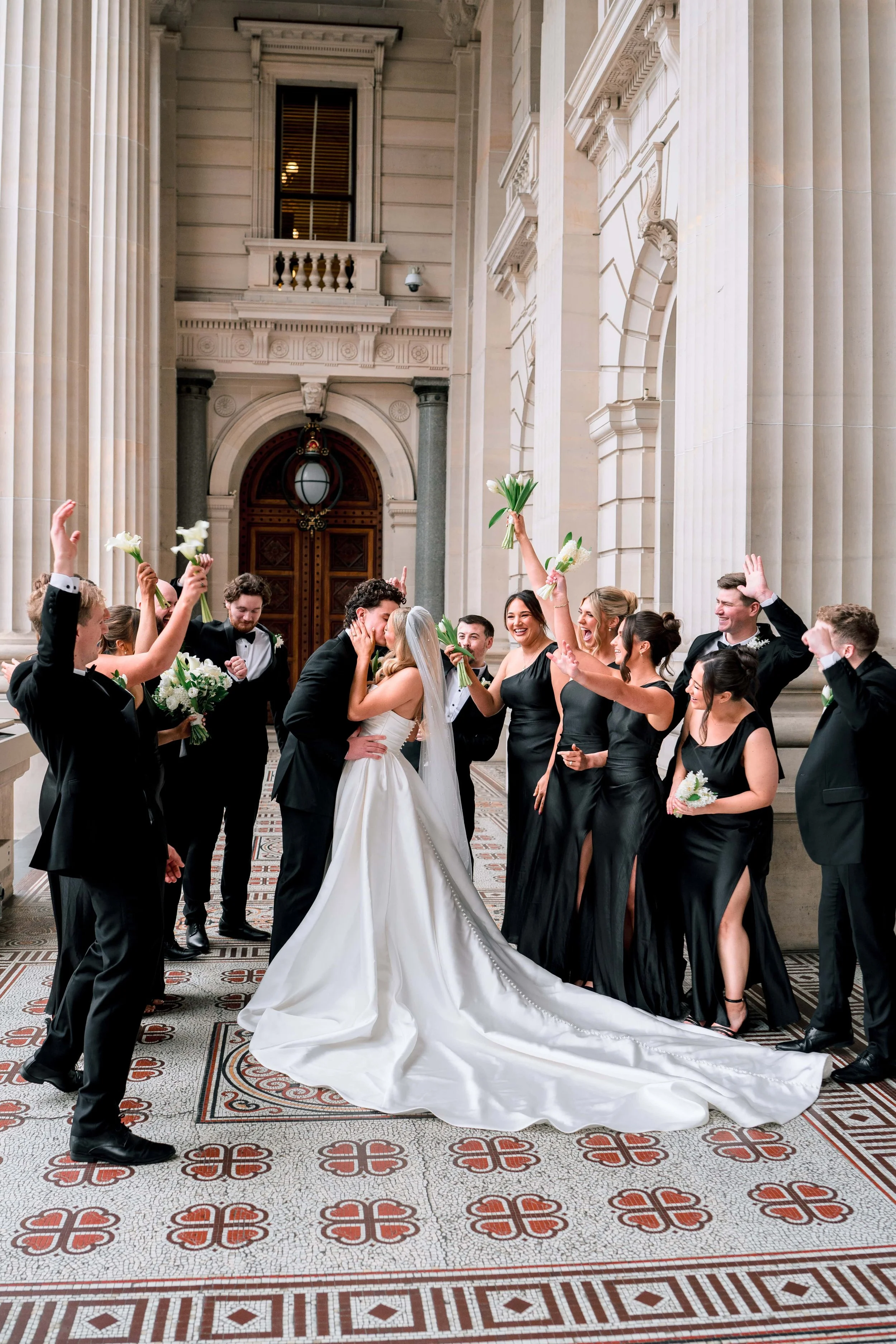 Wedding party inside a grand, historic building with large columns, intricate tile flooring, and high ceilings. The bride and groom are kissing at the center, surrounded by bridesmaids and groomsmen holding bouquets and wearing formal black attire. Some guests are celebrating, raising their hands and flowers in the air.