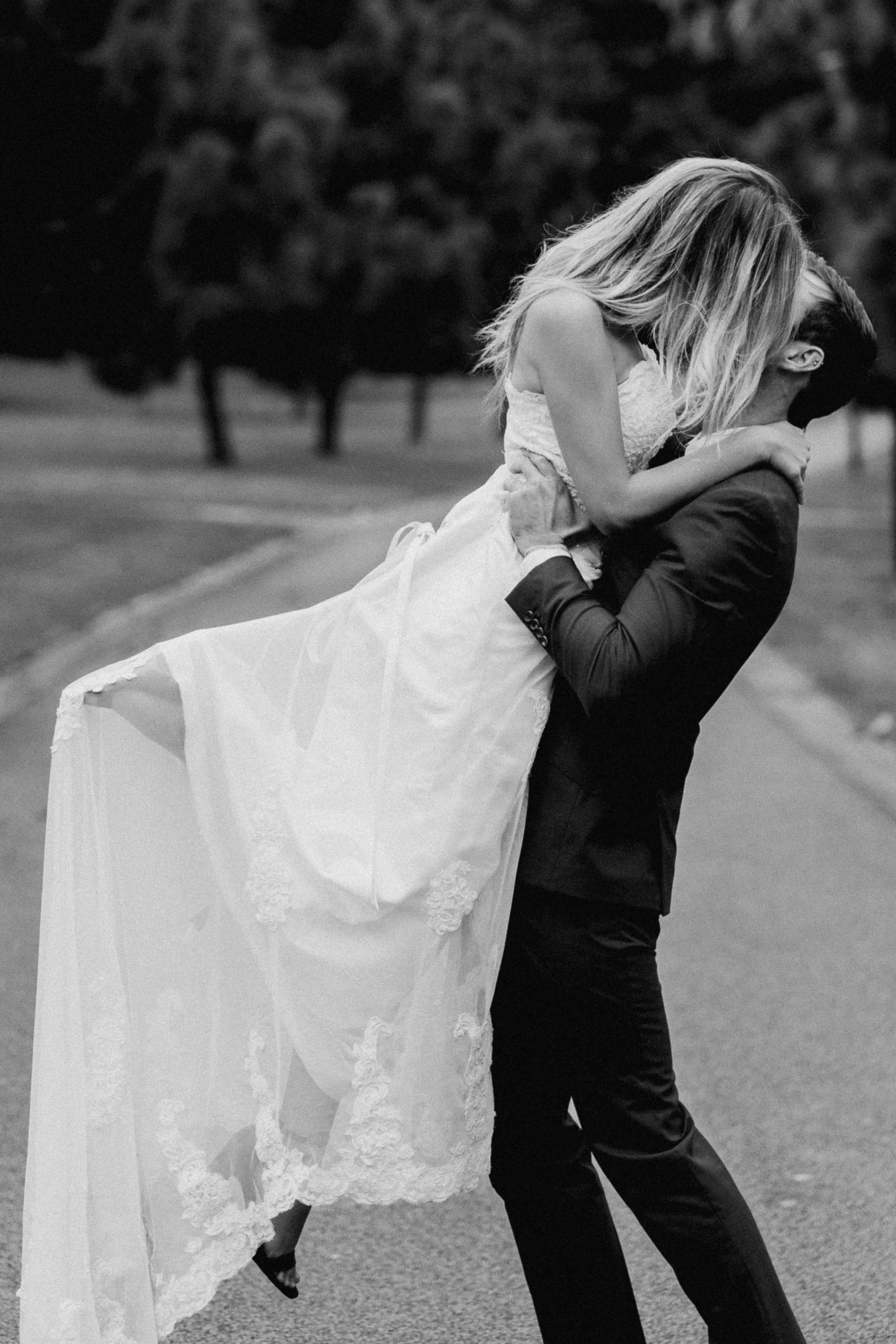 A couple dressed in wedding attire, with the woman wearing a white wedding dress and the man in a dark suit, sharing a kiss outdoors in a park.