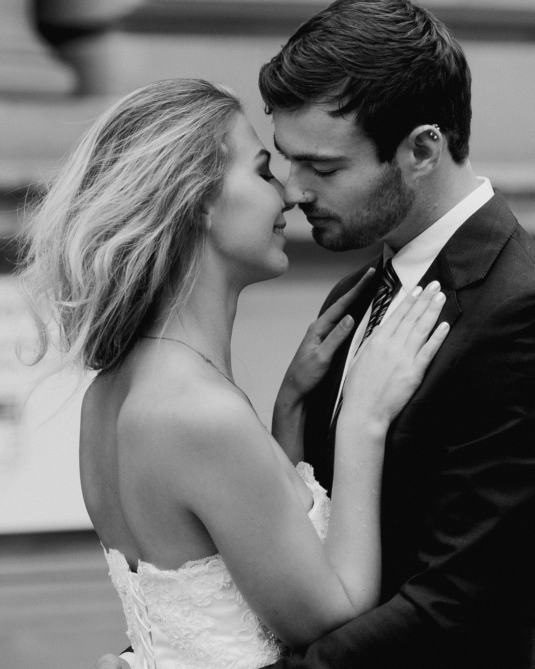 A black-and-white photo of a bride and groom with their foreheads touching, eyes closed, sharing a tender moment during their wedding. The bride is wearing a lace wedding dress, and the groom is in a suit and tie.