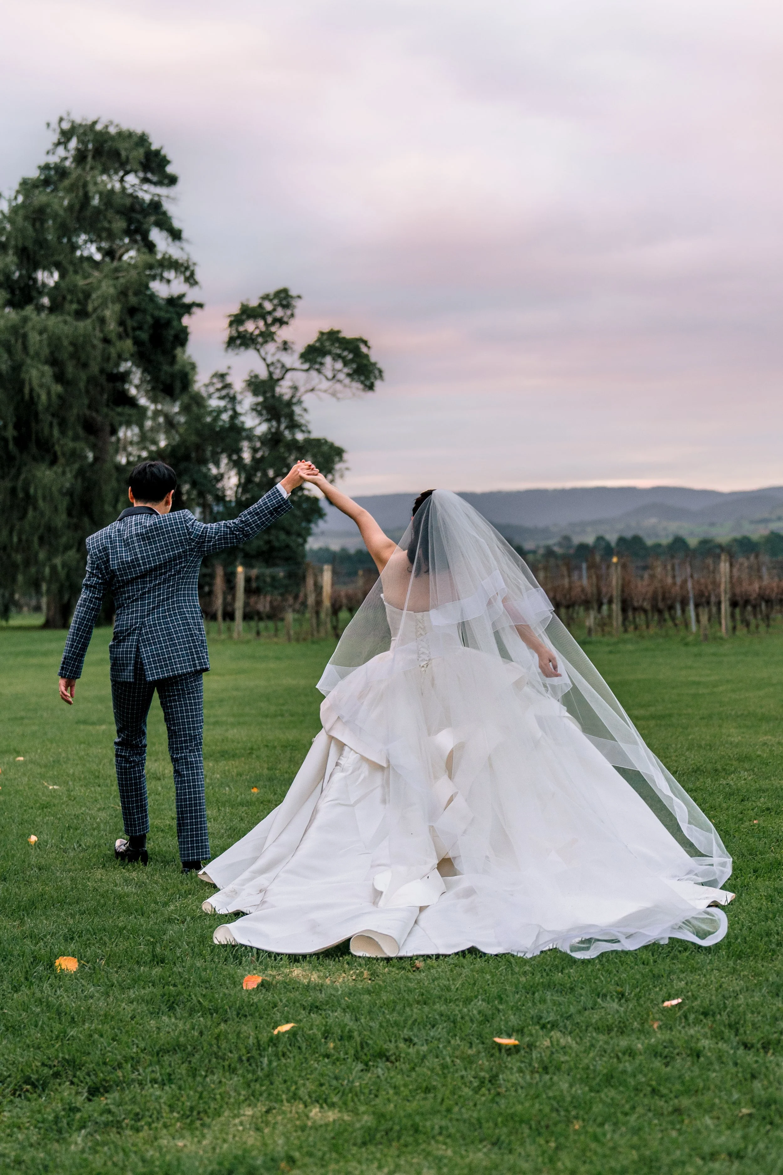 A bride and groom dancing outdoors on a grassy field during a wedding, with trees and mountains in the background, under a cloudy sky.
