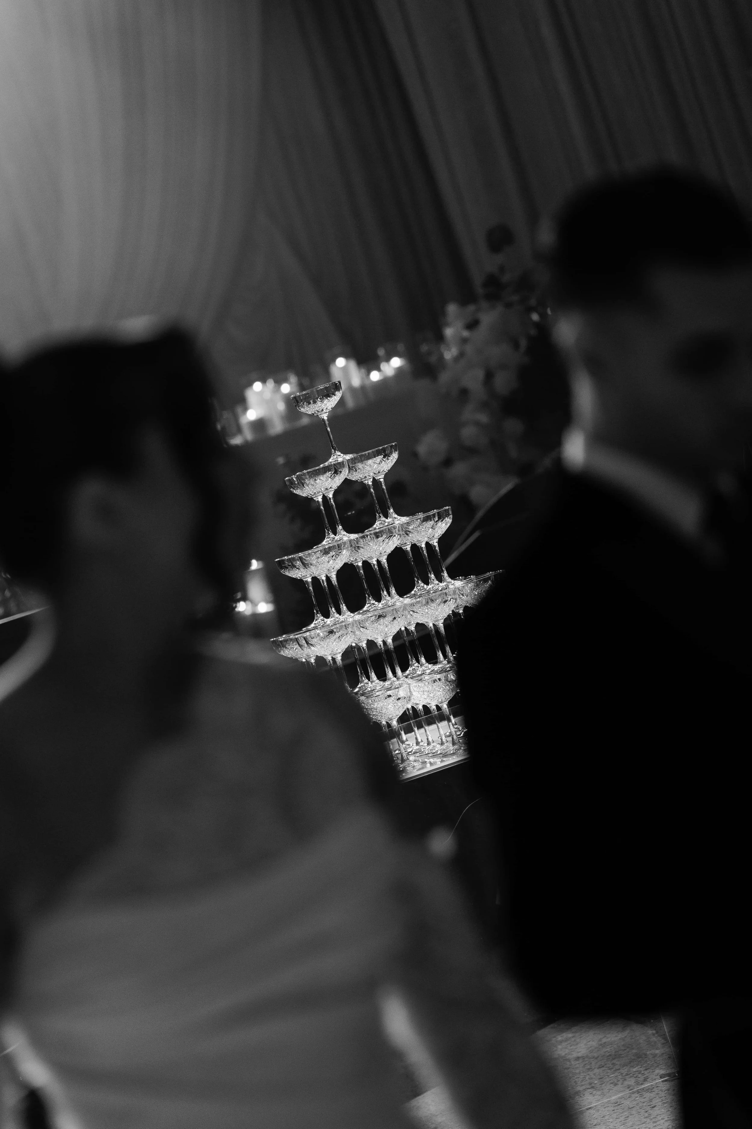 A tiered crystal champagne tower illuminated by ambient lighting, with a blurred background of candles and floral decorations at a celebration or event.