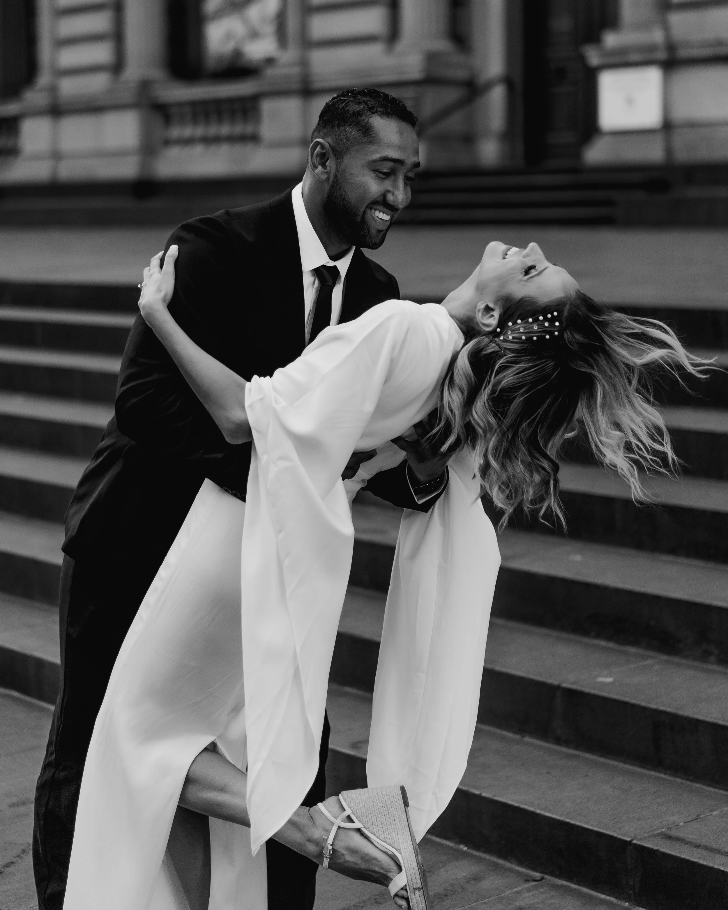 A man in a suit and a woman in a dress are dancing together, smiling and looking at each other, on a staircase inside a building.