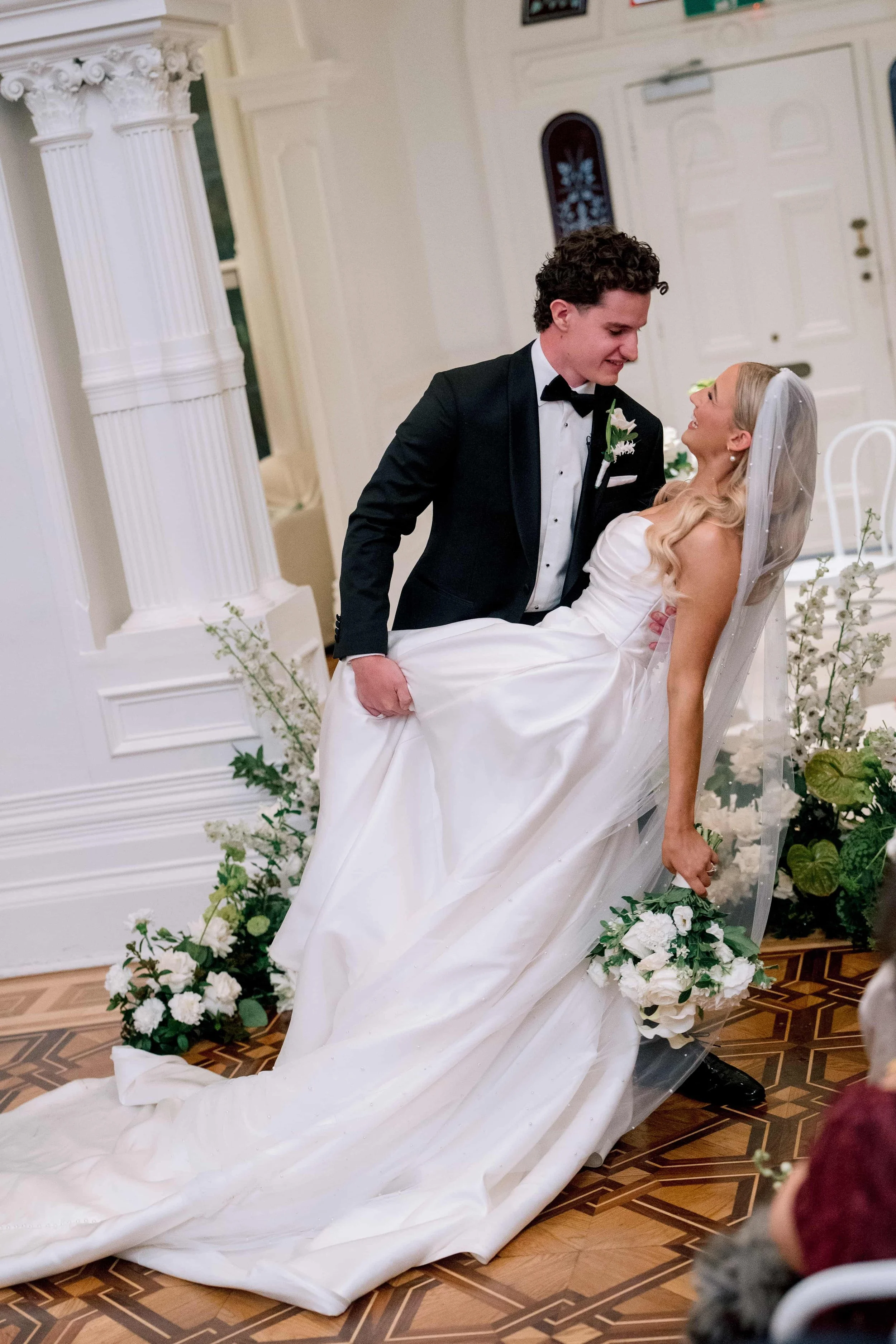 A bride and groom sharing a moment during their wedding ceremony. The groom is standing and smiling down at the bride, who is seated in a chair, looking up at him with a joyful expression. The bride is wearing a white wedding gown and veil, and the groom is dressed in a tuxedo with a bow tie. The setting is decorated with white flowers and greenery.