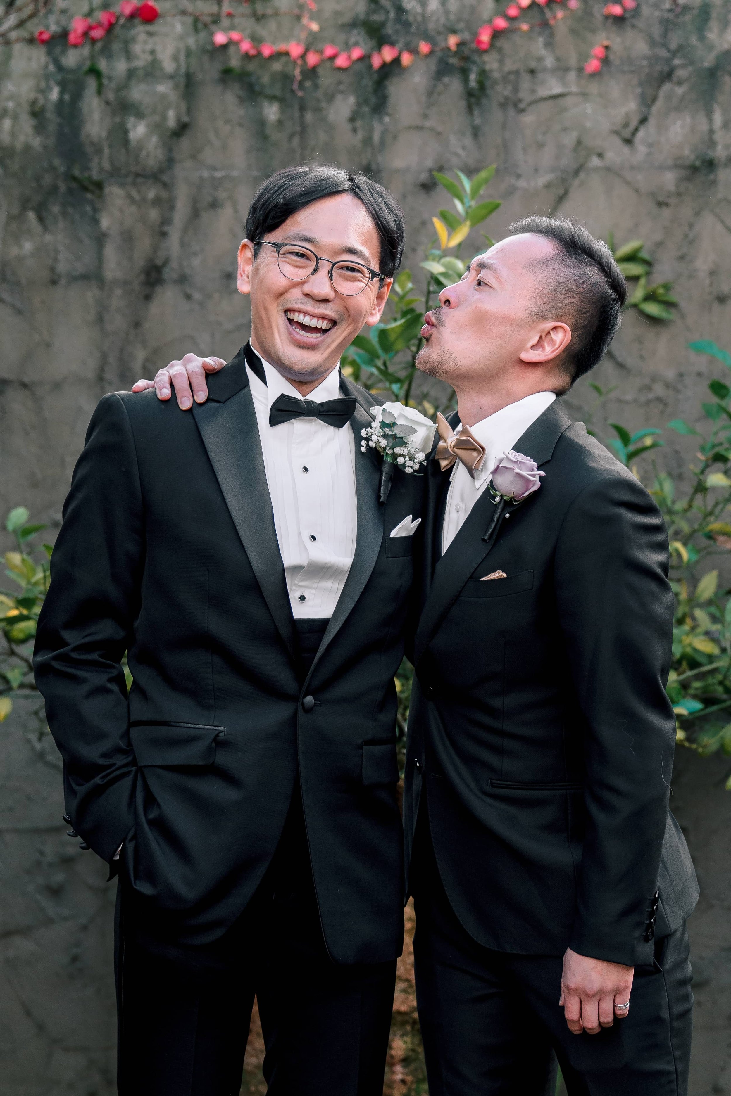 Two men in tuxedos share a joyful moment outdoors, one with his arm around the other's shoulder, against a background of a stone wall with plants.