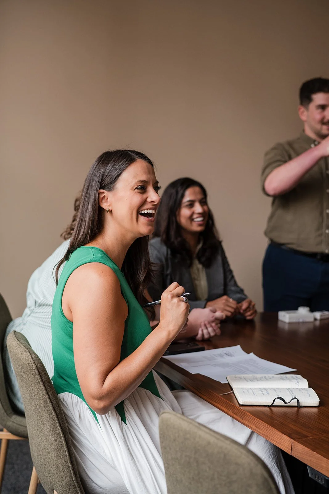 Group of people sitting at a conference table, smiling and engaging in a discussion. Notebooks, papers, and pens are visible on the table.
