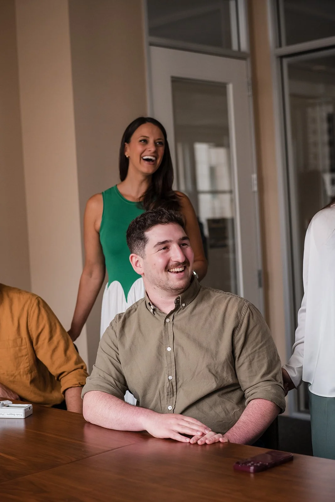 A group of people sitting at a conference table, smiling and laughing while a woman in a green and white dress stands behind them.