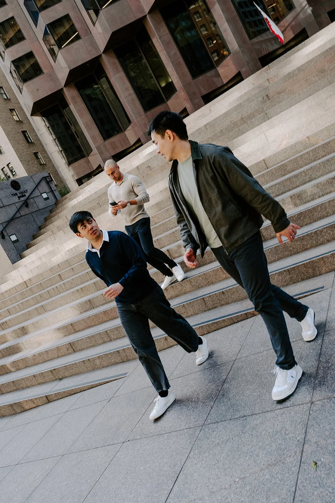 Three young men walking down concrete steps outside a modern glass building, one looking at his phone, and the others engaged in conversation.